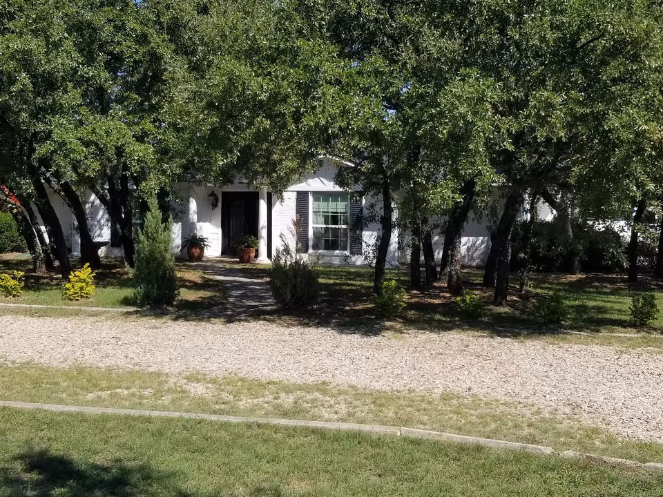 Exterior of a white senior living facility front partially obscured by trees and a gravel path.