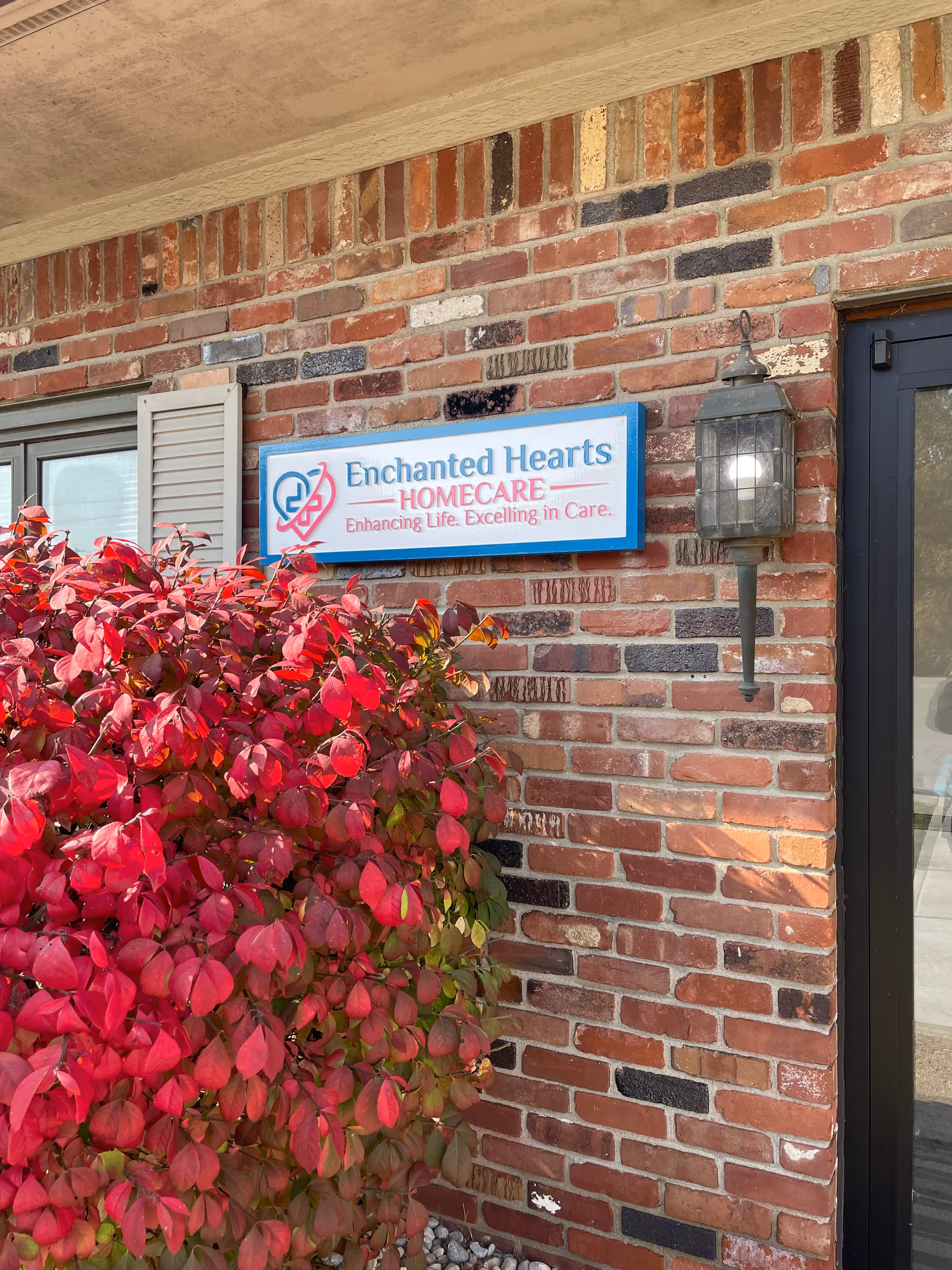 Brick exterior with Enchanted Hearts Homecare sign, lantern, and red flowering shrub.