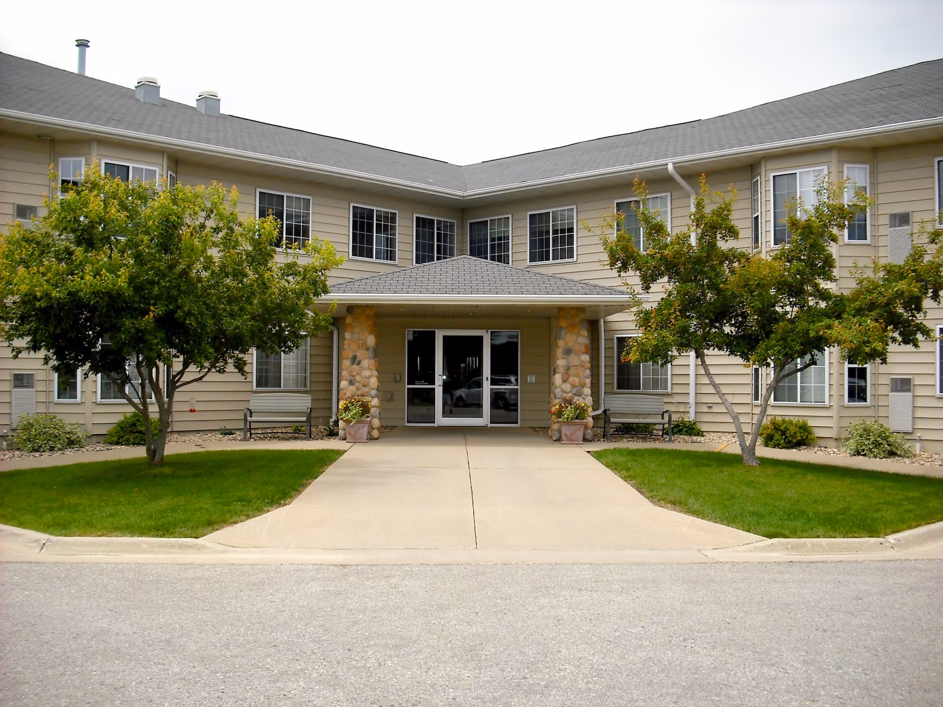 Exterior view of a beige senior living building with entrance, benches, and trees.