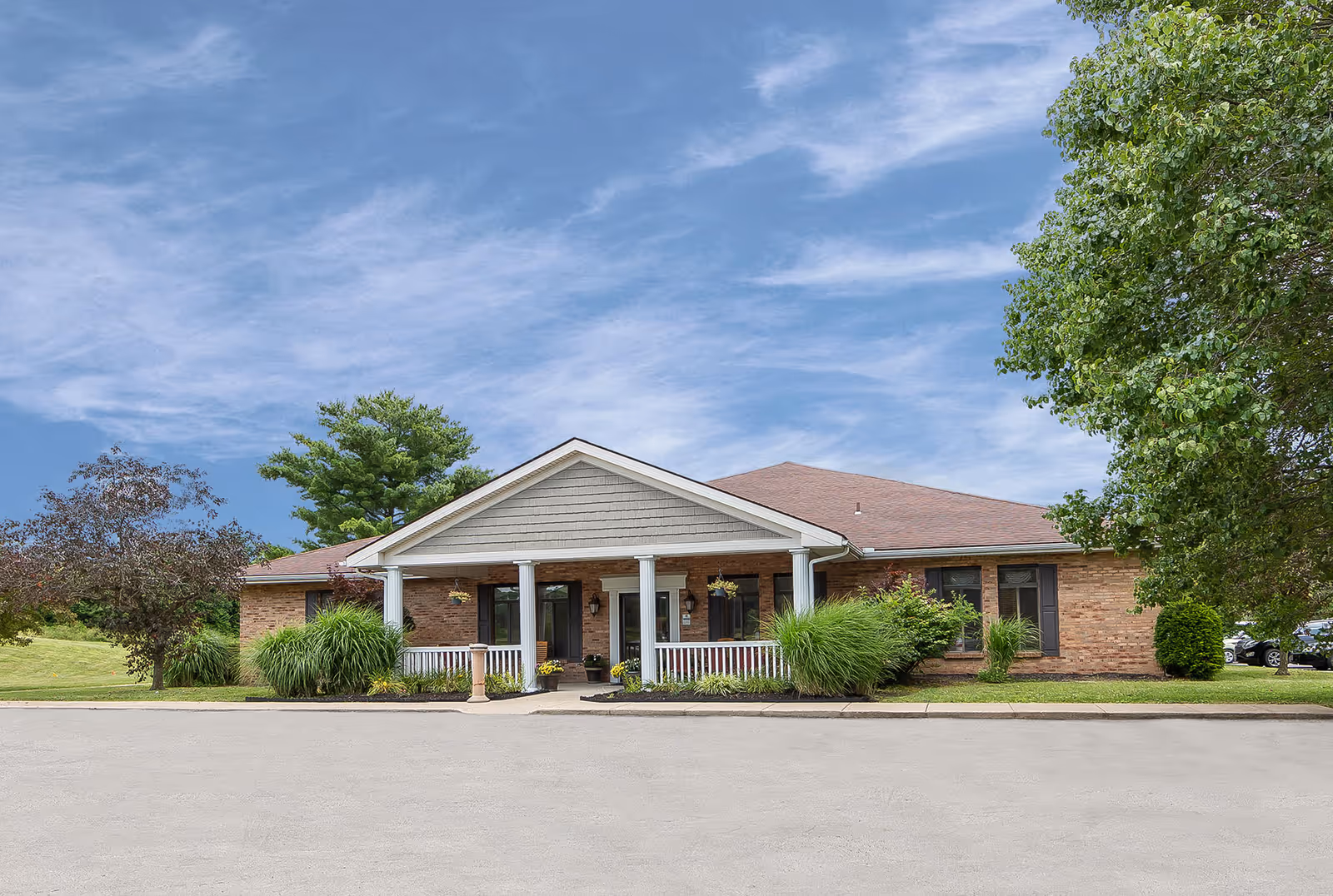 Front exterior view of a single-story brick building with a covered entrance supported by white columns, surrounded by green shrubs and trees under a partly cloudy blue sky.