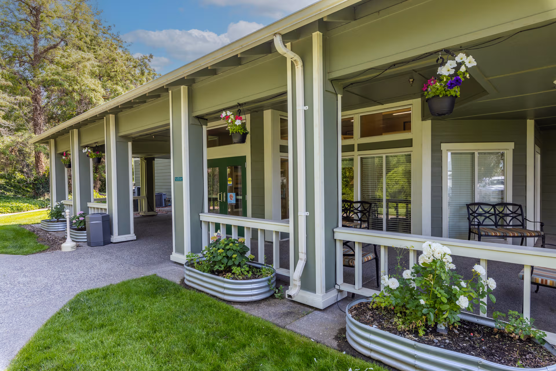 Green-painted assisted living building with a covered front porch, planters, and hanging baskets.