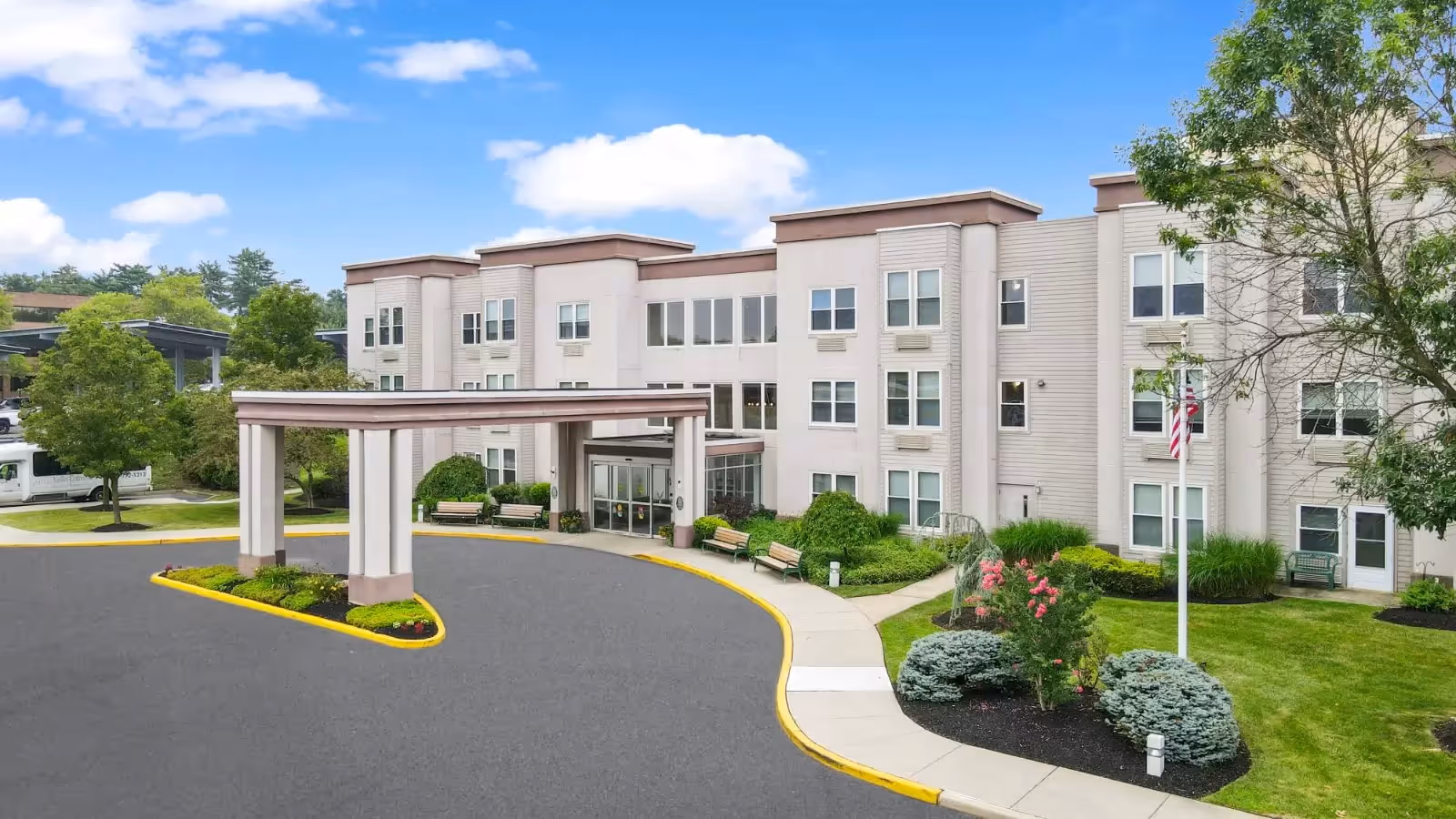 Exterior view of a three-story senior living facility building with beige siding and multiple windows. The entrance features a covered drop-off area with benches nearby. The surrounding area includes well-maintained landscaping with green grass, bushes, trees, and a flagpole with an American flag. The sky is partly cloudy.