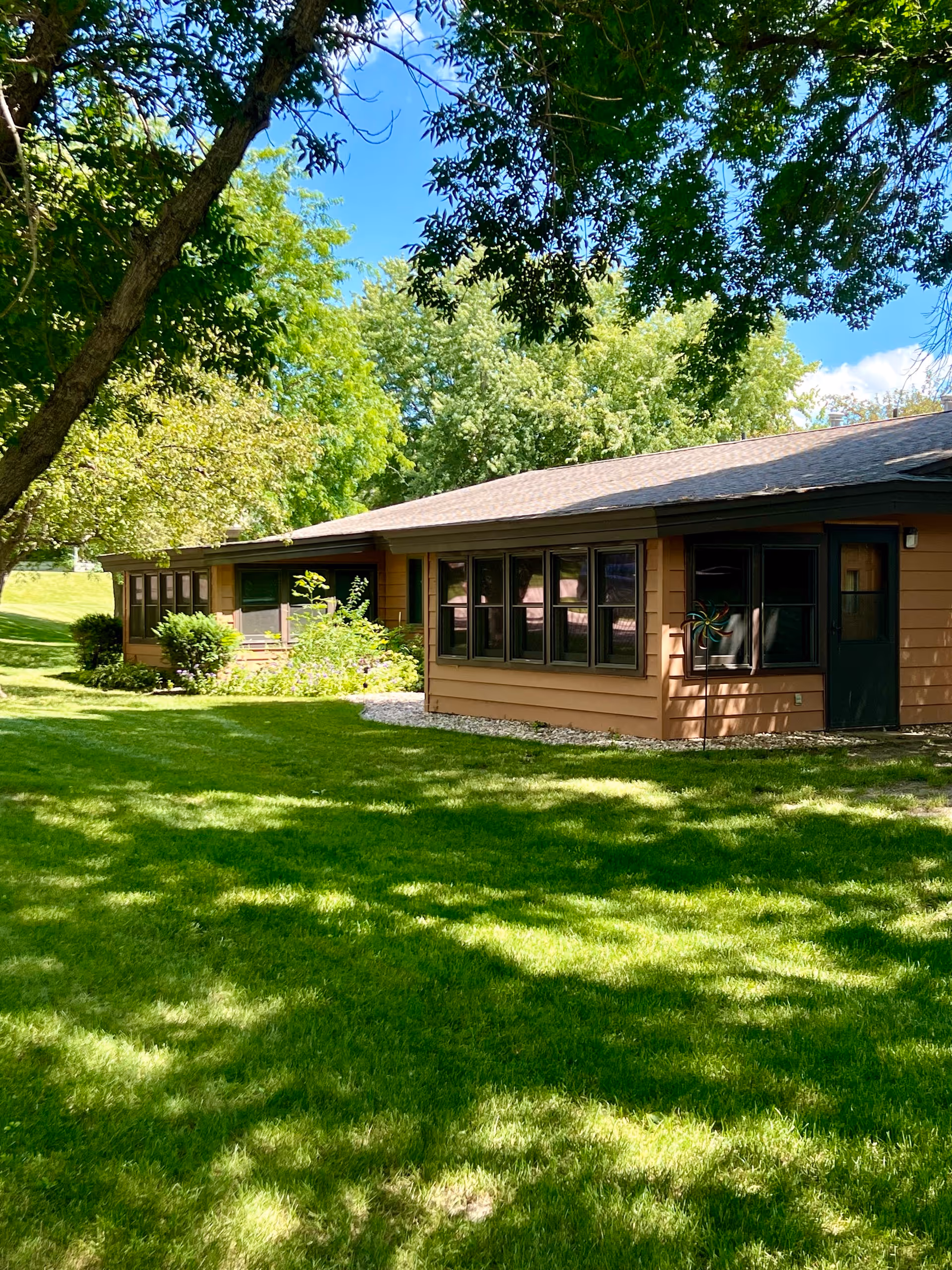 A single-story brown building with multiple windows surrounded by lush green grass and trees under a clear blue sky, casting shadows on the lawn.