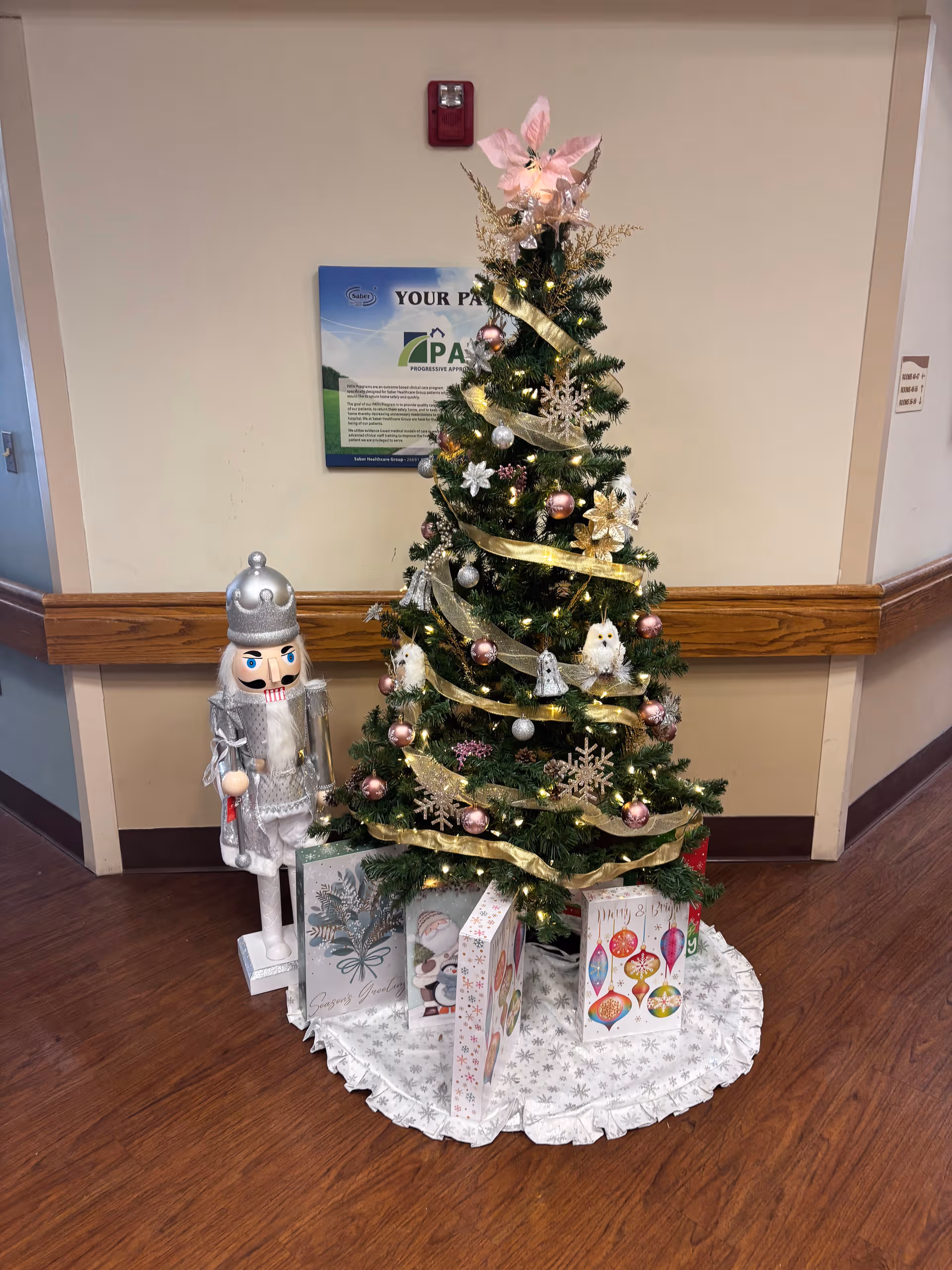 Holiday-decorated hallway with Christmas tree, gifts, and a nutcracker display in a senior facility.