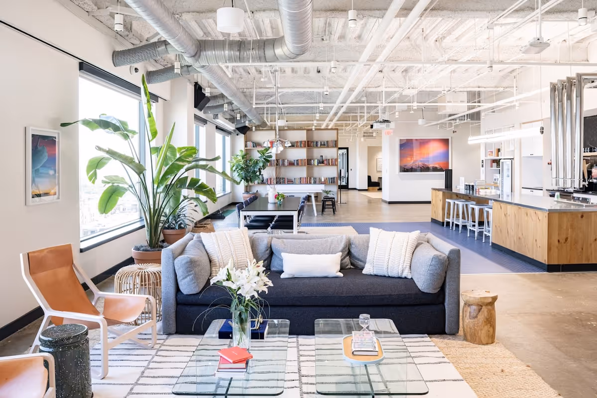 Open-concept lounge in a modern facility with sofa, plants, bookshelves, and a kitchen.