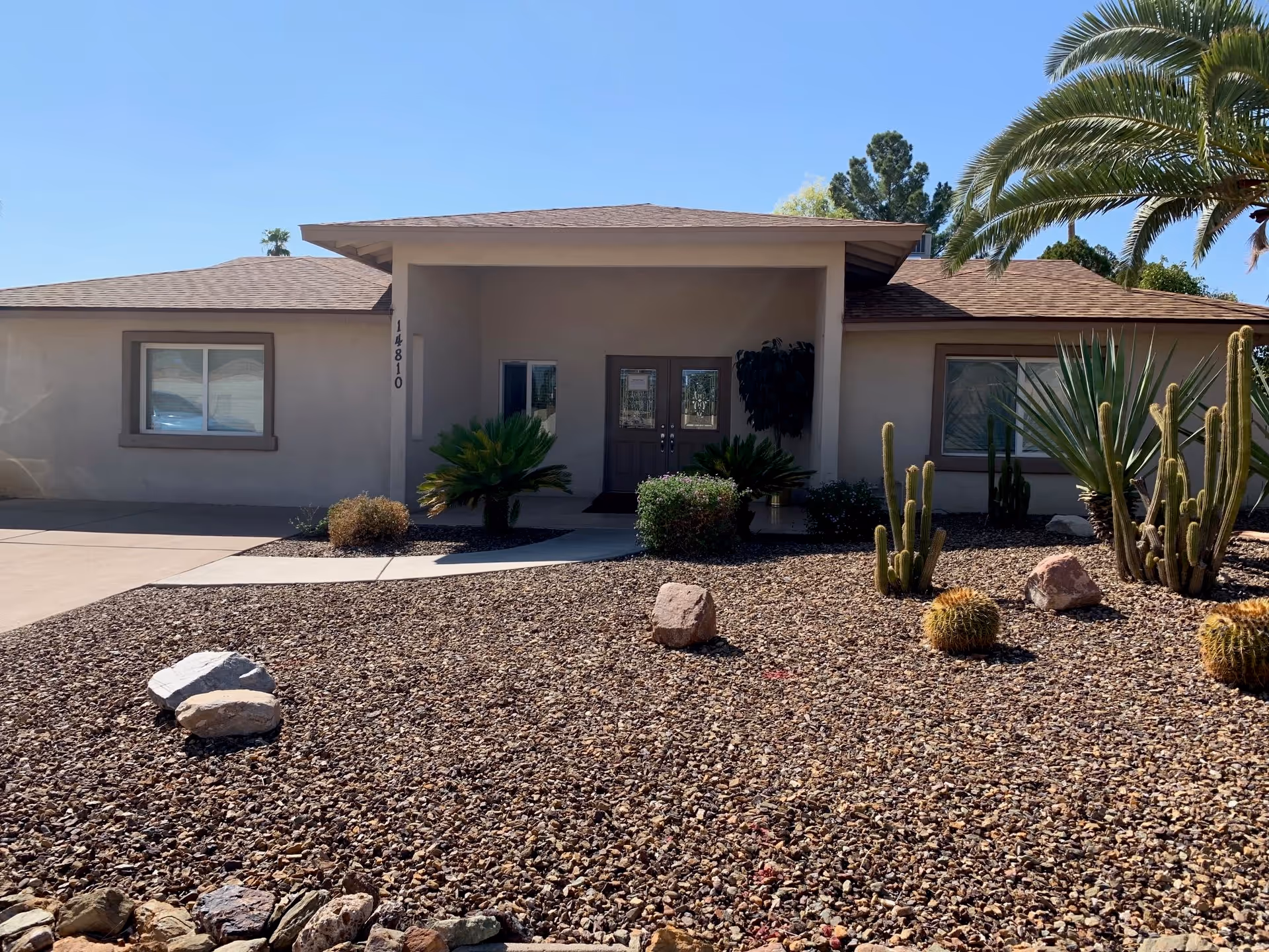 Front exterior view of a single-story building with a covered entrance, desert landscaping with rocks, cacti, and palm trees, and a clear blue sky.