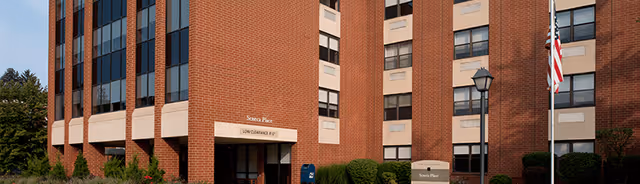 Front exterior of a brick multi-story building labeled 'Seneca Place' with an entrance canopy, flagpole, mailbox, and landscaping.