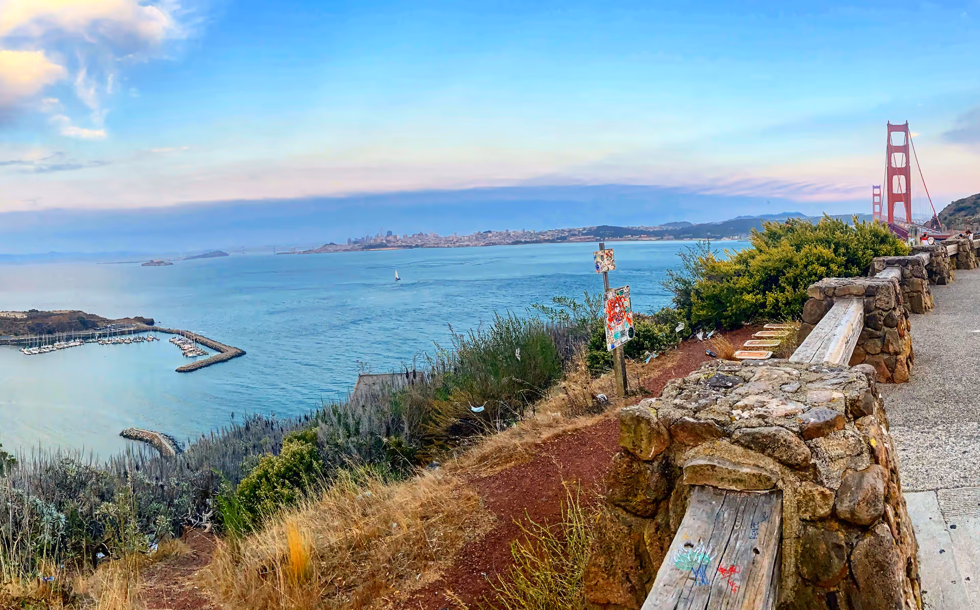 Scenic coastal overlook with a stone wall, benches, and a distant red bridge over the water.