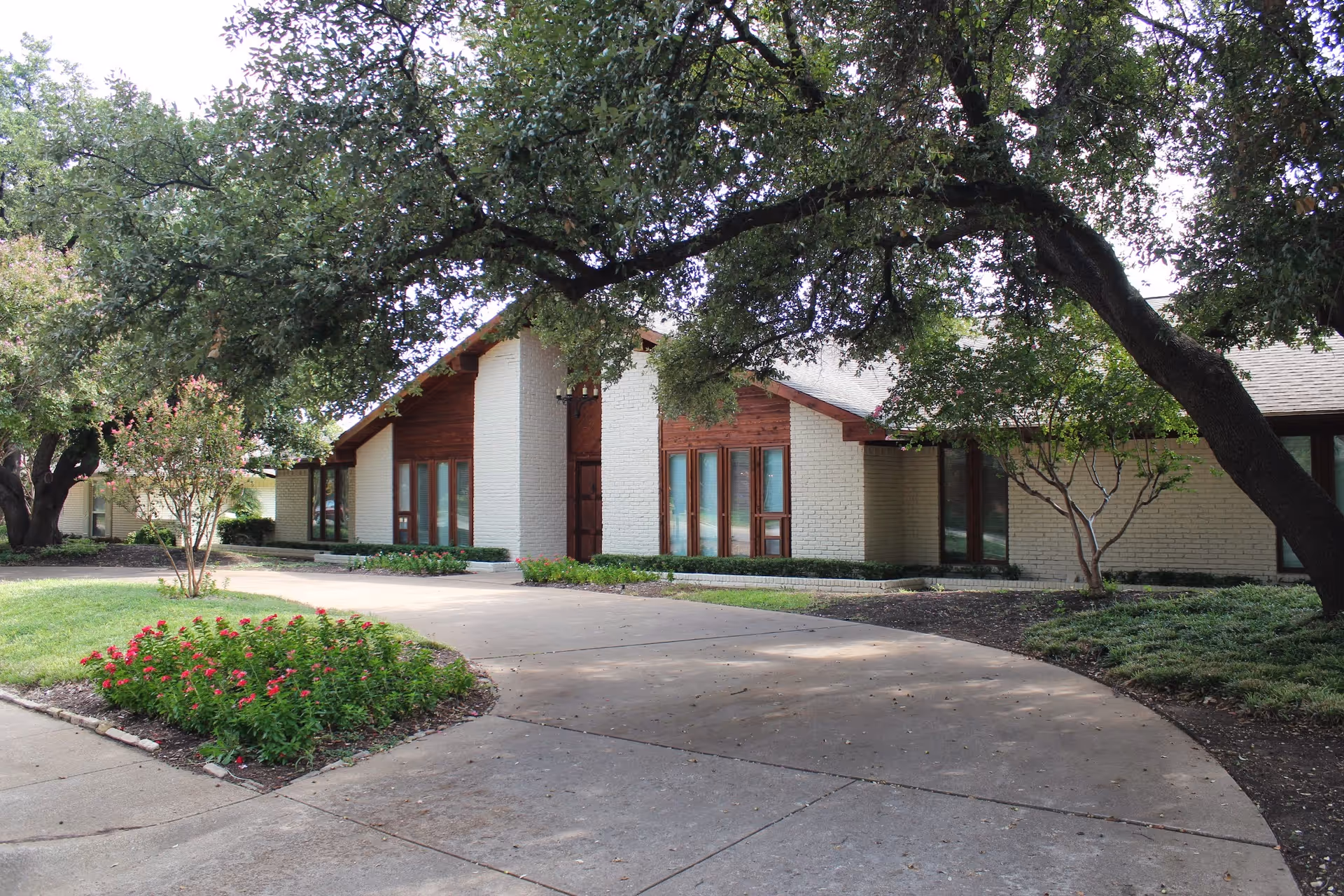 Exterior view of a single-story building with white brick walls and wooden accents, surrounded by large trees and landscaped greenery including a flower bed with red flowers. A curved concrete driveway leads to the entrance.