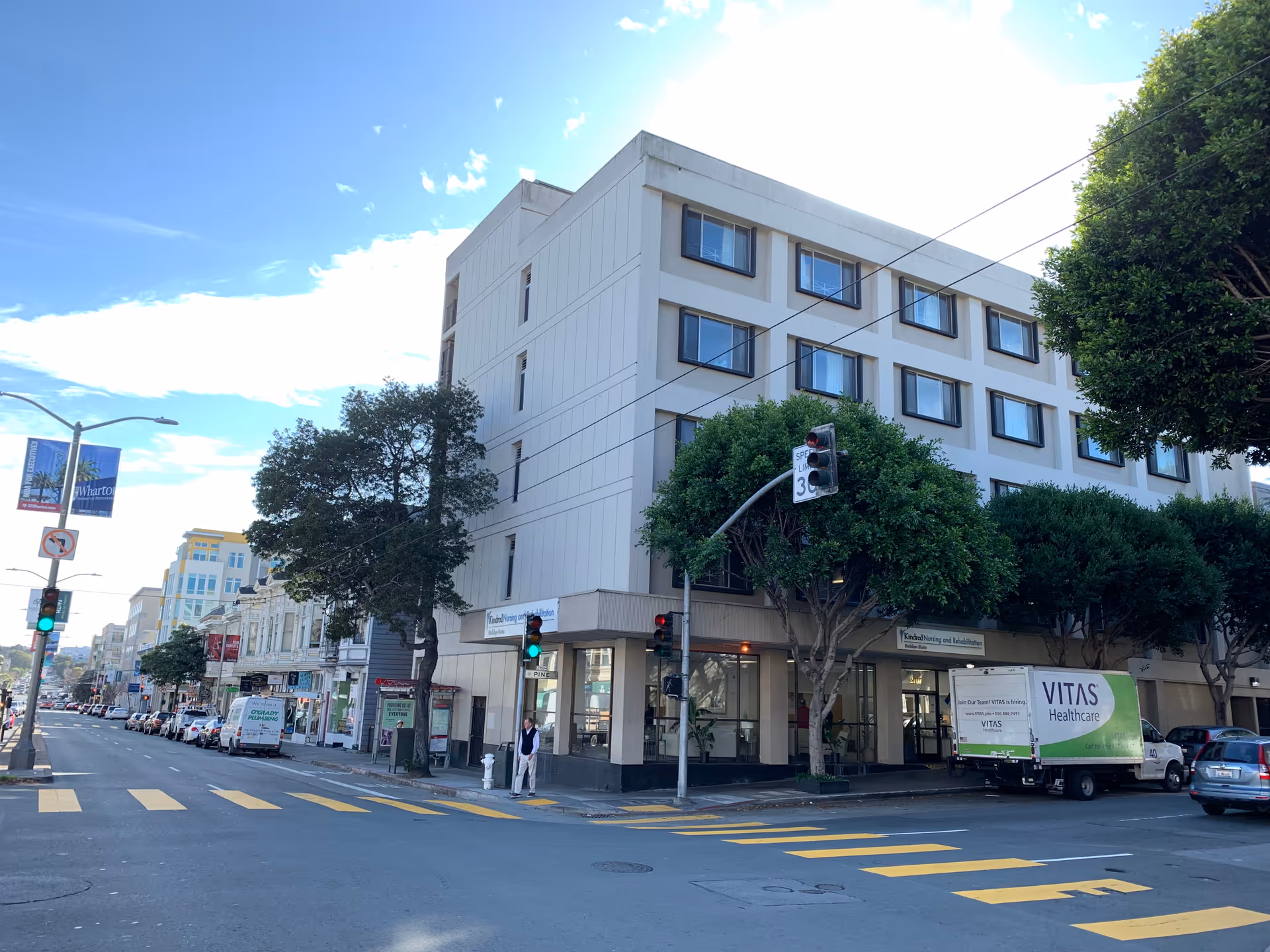Street view of a multi-story building on a sunny day with trees lining the sidewalk. The building has multiple windows and a sign for Kindred Nursing and Rehabilitation. A VITAS Healthcare truck is parked near the entrance. Traffic lights and street signs are visible, along with a pedestrian standing on the sidewalk.