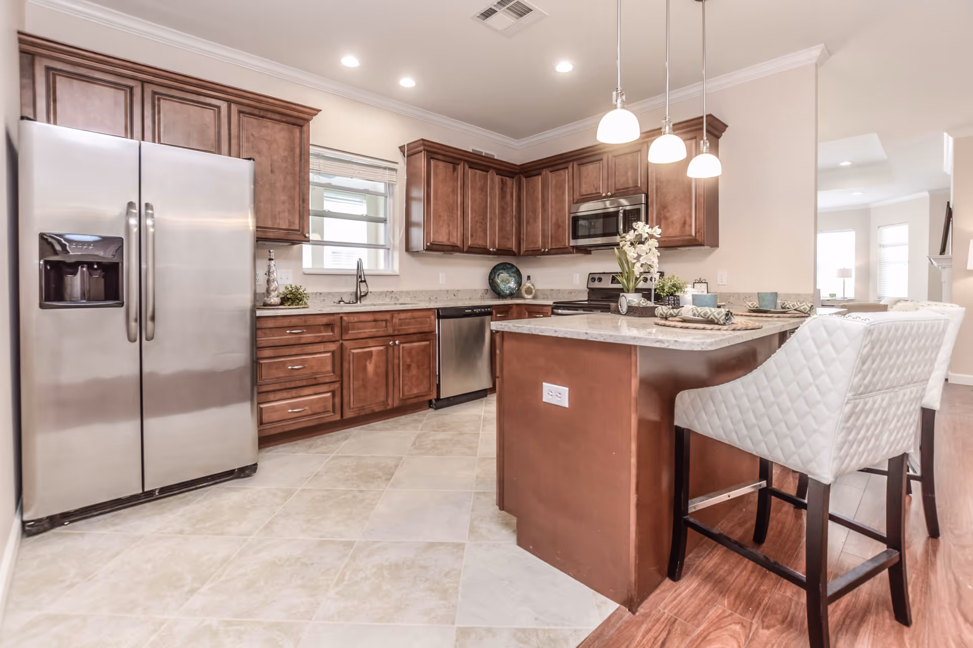 Open modern kitchen with stainless steel appliances, wooden cabinets, a central island with pendant lights and upholstered bar stools.