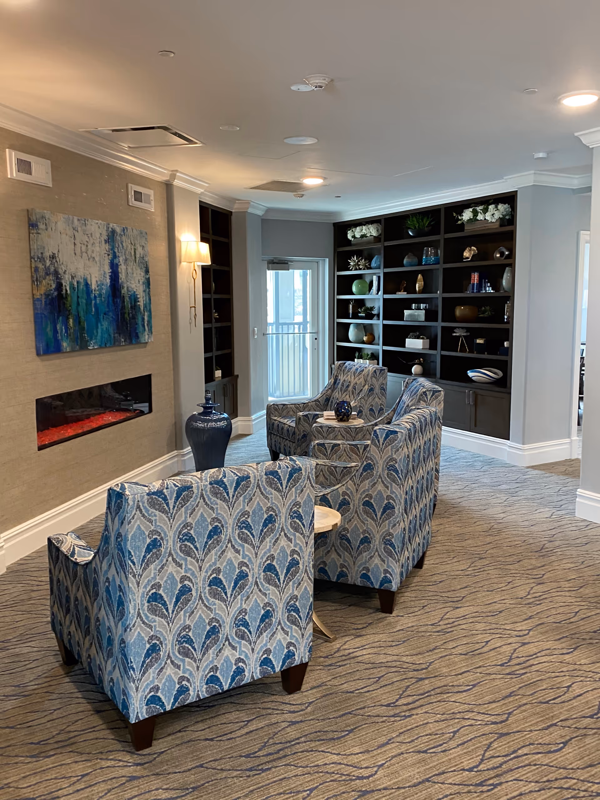 Senior living facility lounge with blue patterned chairs, shelves, and abstract artwork.