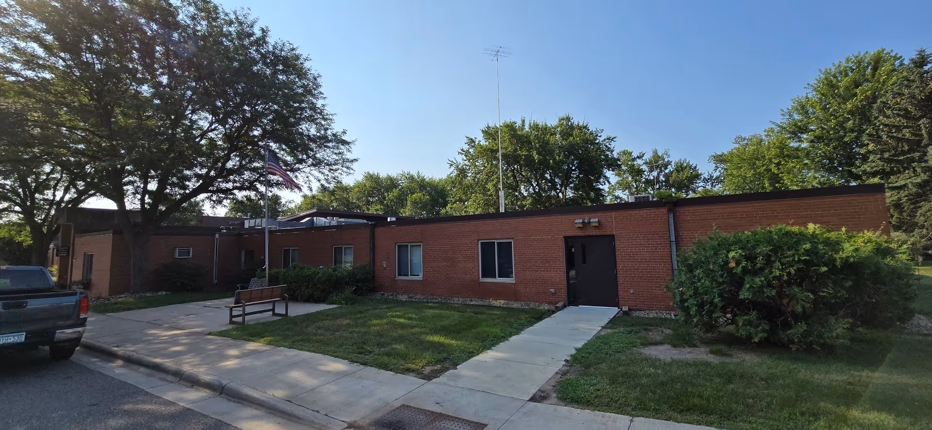 Brick, single-story senior living building with a walkway, bench, flagpole, and trees.