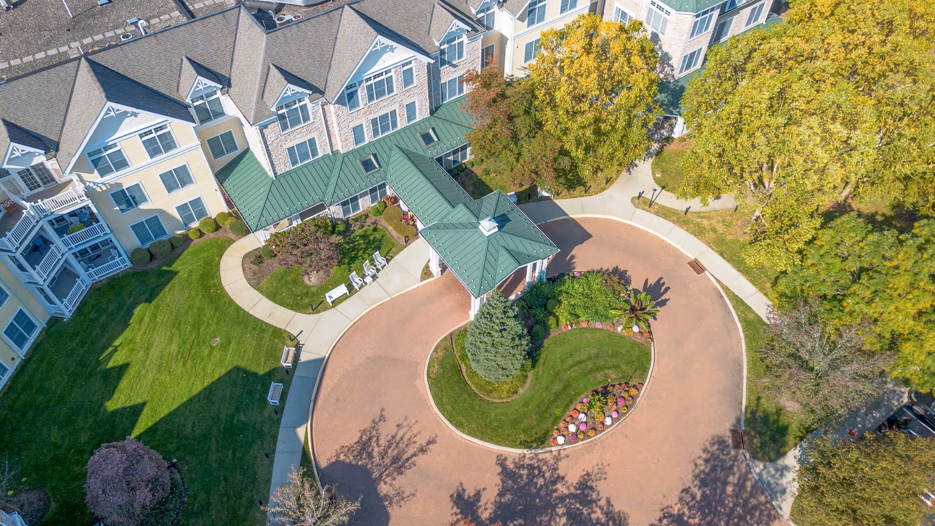 Aerial view of the entrance area of a senior living facility with a circular driveway, landscaped garden with flowers and trees, and a covered drop-off area. The building has multiple stories with beige and stone exterior walls and green roofs.