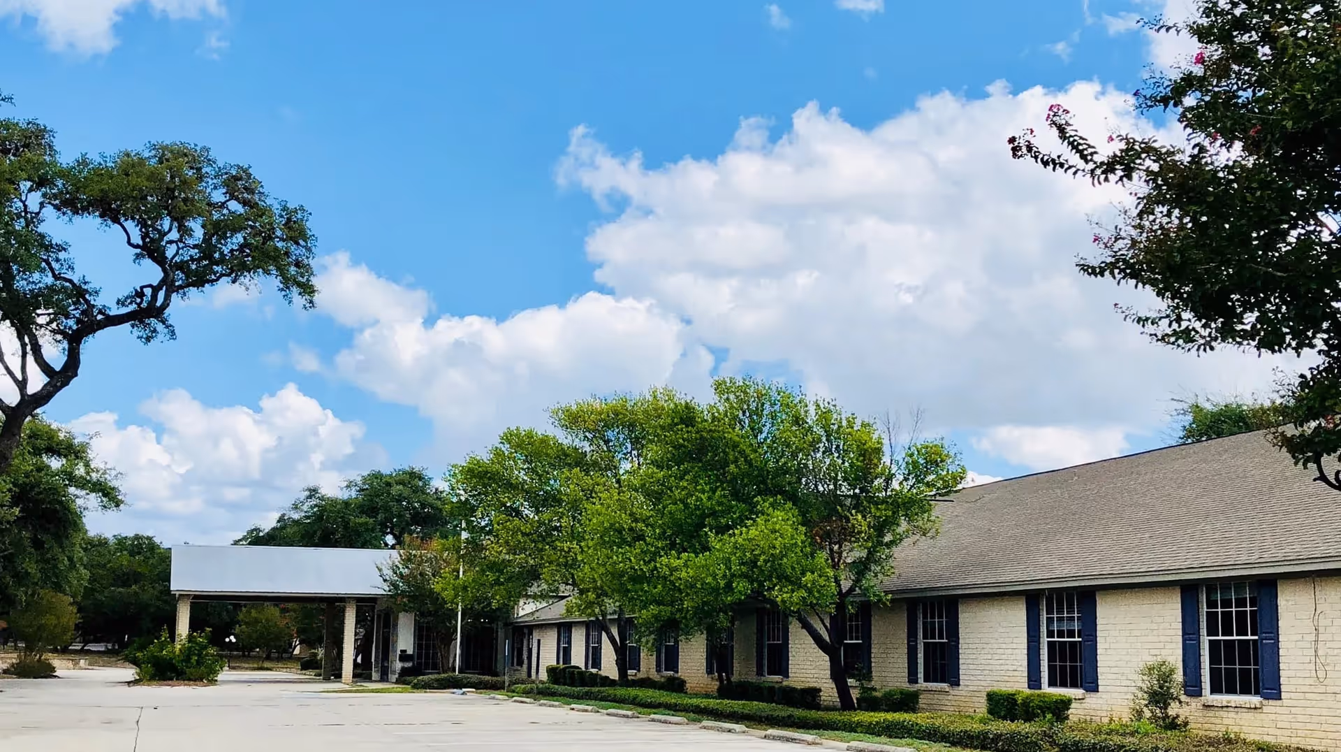 Exterior view of a single-story brick building with blue window shutters, surrounded by green trees and bushes under a partly cloudy blue sky. There is a covered entrance and a large paved parking area in front.
