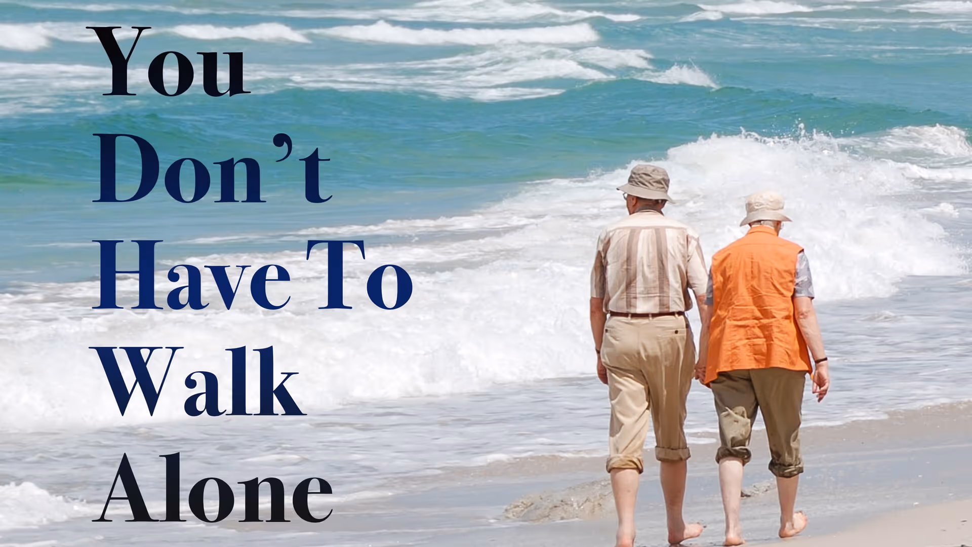 Two elderly people walk barefoot along a sandy beach with waves rolling in.