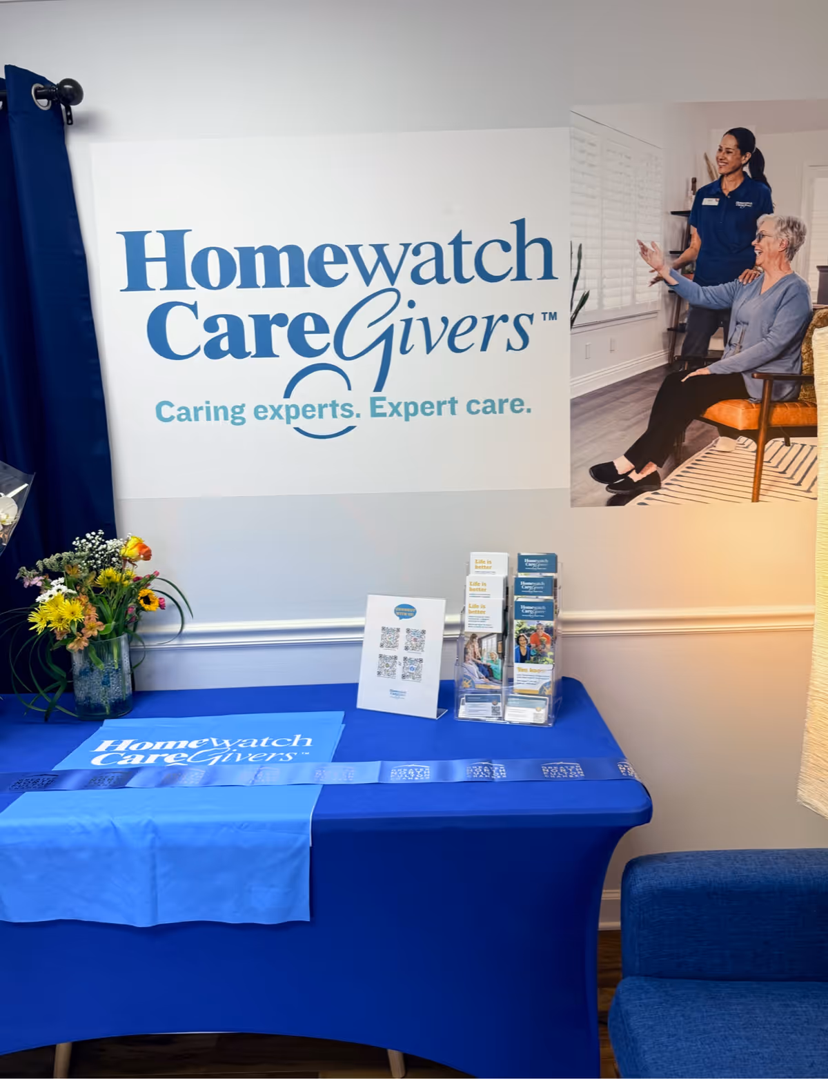 Interior display area featuring Homewatch CareGivers sign, blue table, brochures, and a vase of flowers.