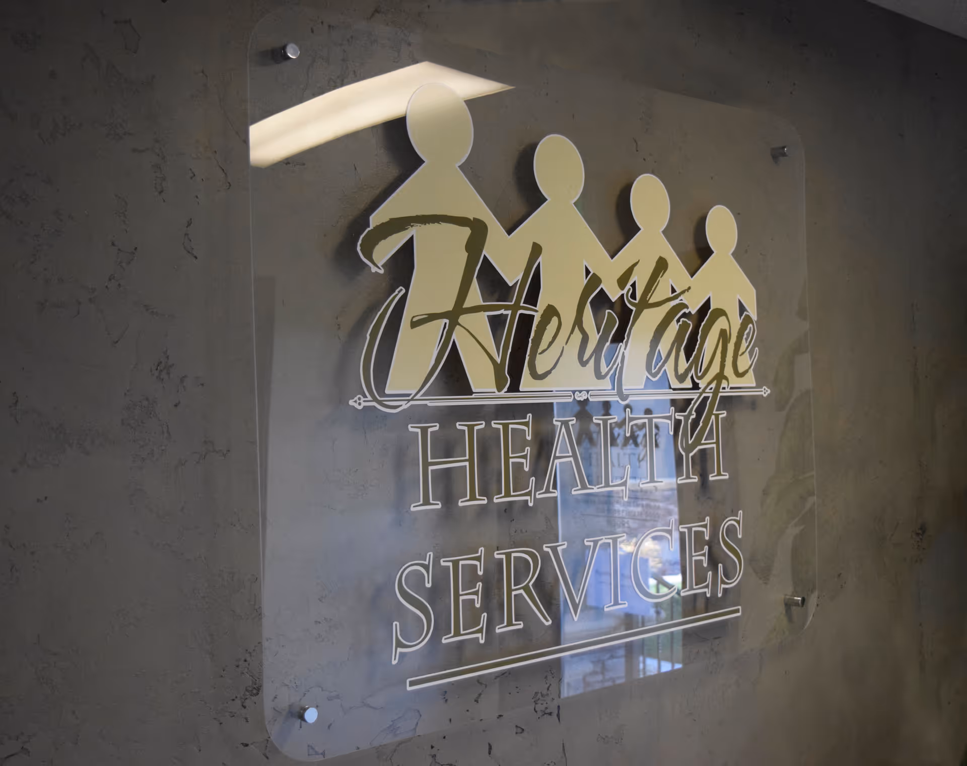 Clear acrylic sign on textured wall showing Heritage Health Services logo with a family icon