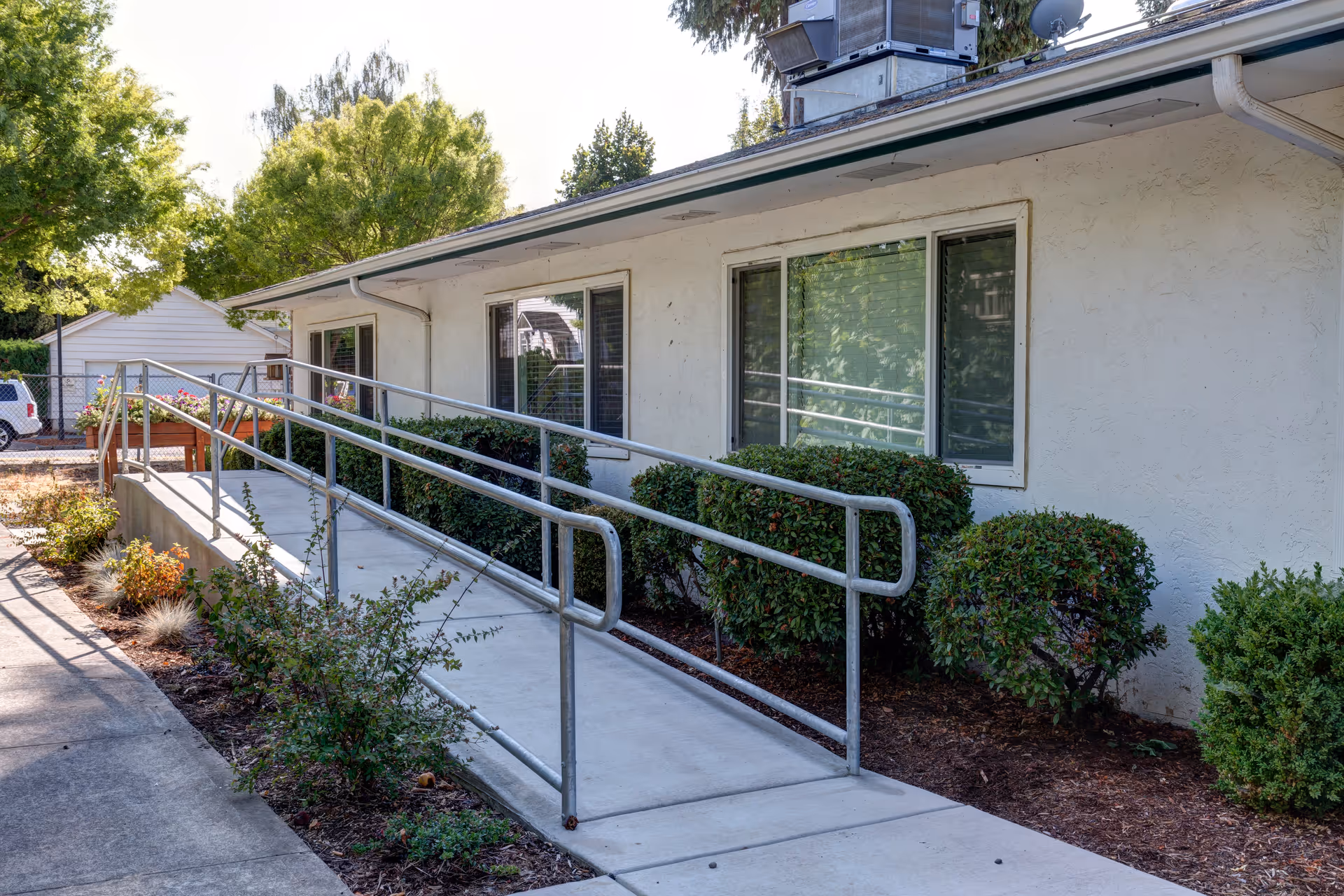 Exterior view of a single-story building with a concrete wheelchair ramp and metal handrails leading to the entrance. The building has white stucco walls and several windows with blinds. There are bushes and small plants along the side of the ramp, and trees in the background under a clear sky.