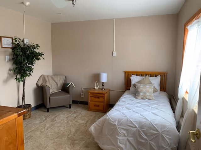 Tidy bedroom with a single bed, nightstand, lamp, chair, plant, and window curtains.