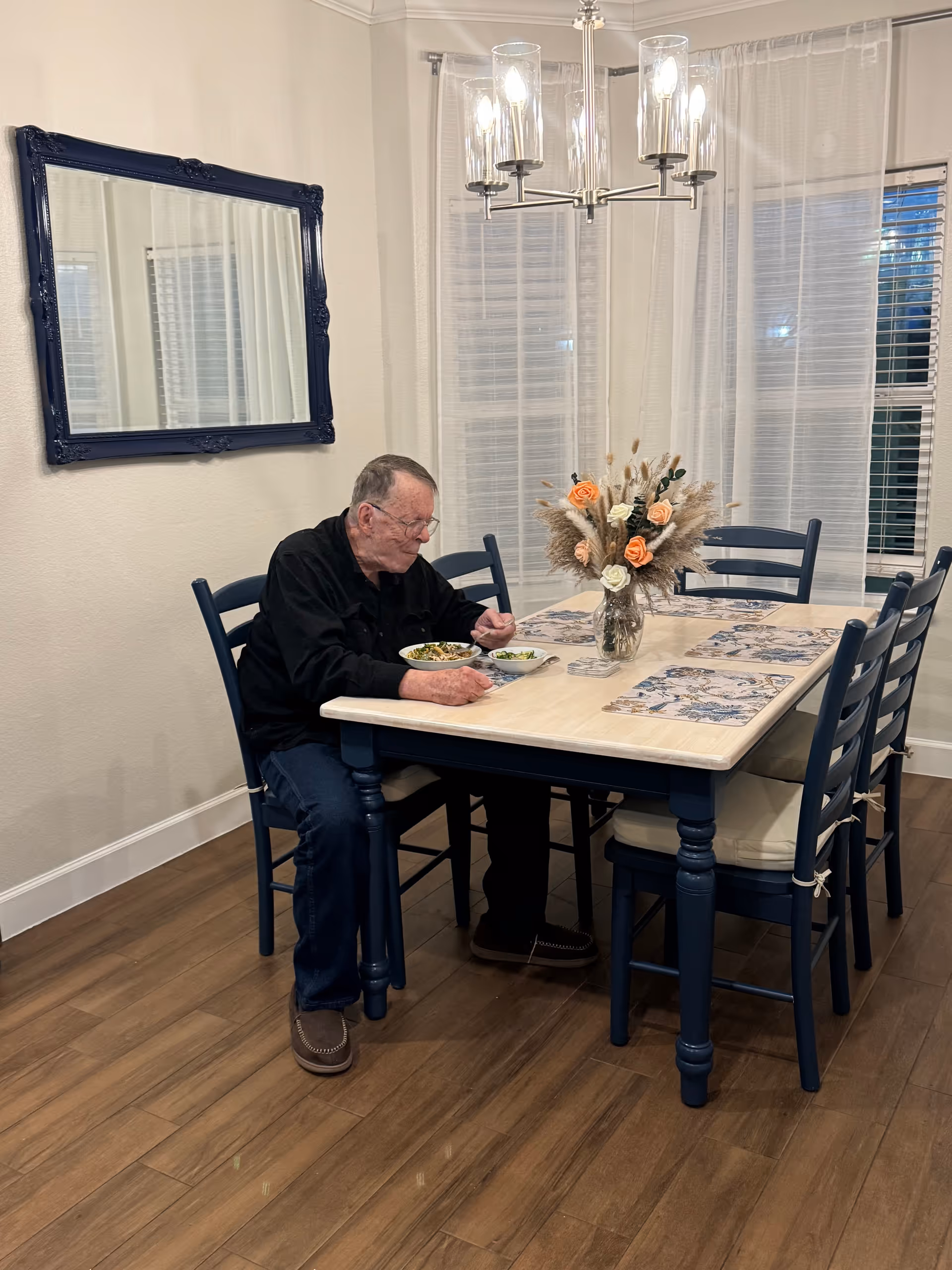 Elderly man seated at a dining table eating a meal in a bright dining room.