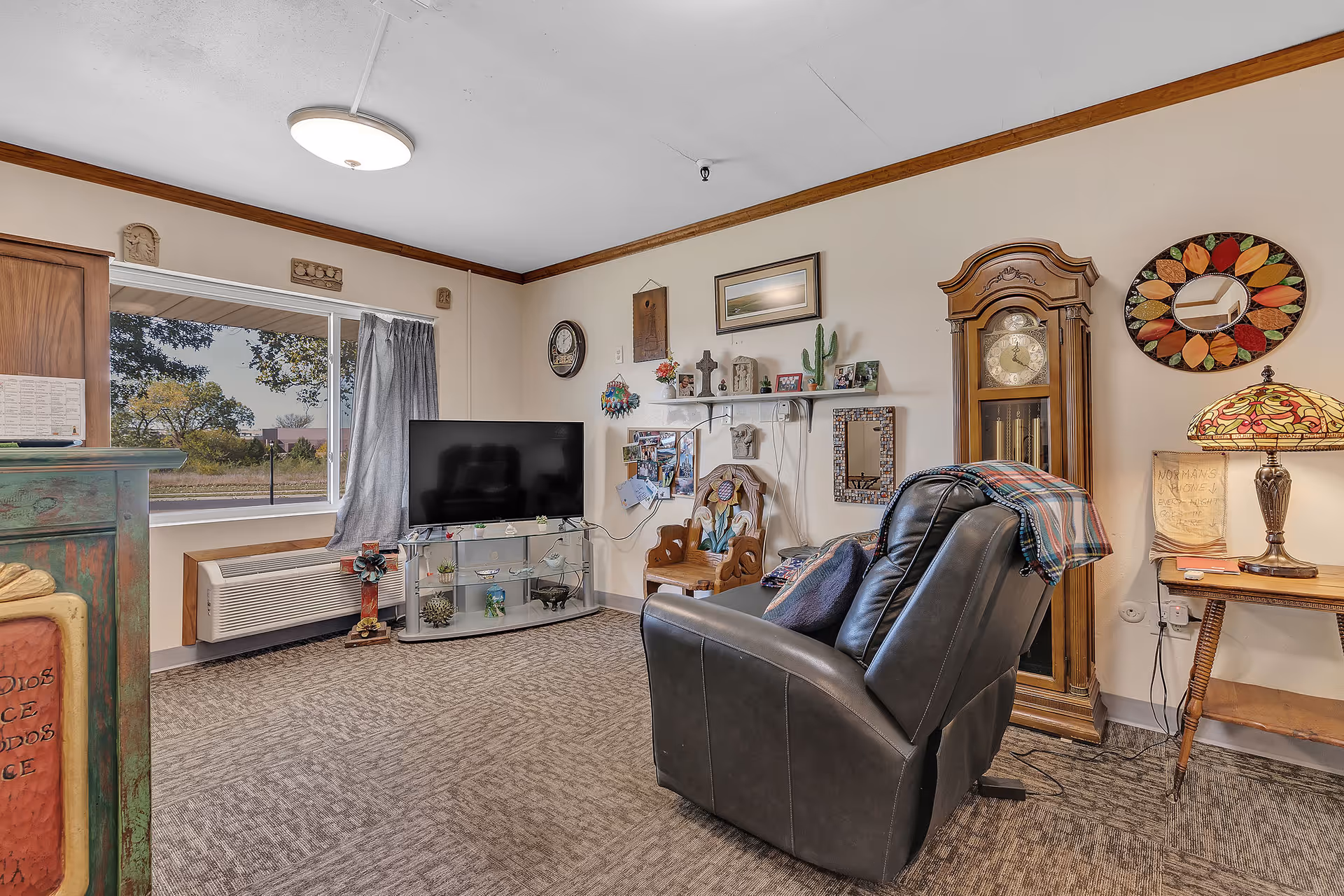 A cozy living room area in a senior care facility featuring a black recliner chair with a colorful blanket draped over it, a grandfather clock, a TV on a glass stand, various wall decorations including a round mirror with a colorful leaf pattern, and a Tiffany-style table lamp on a wooden side table. A large window with gray curtains lets in natural light and shows trees outside.