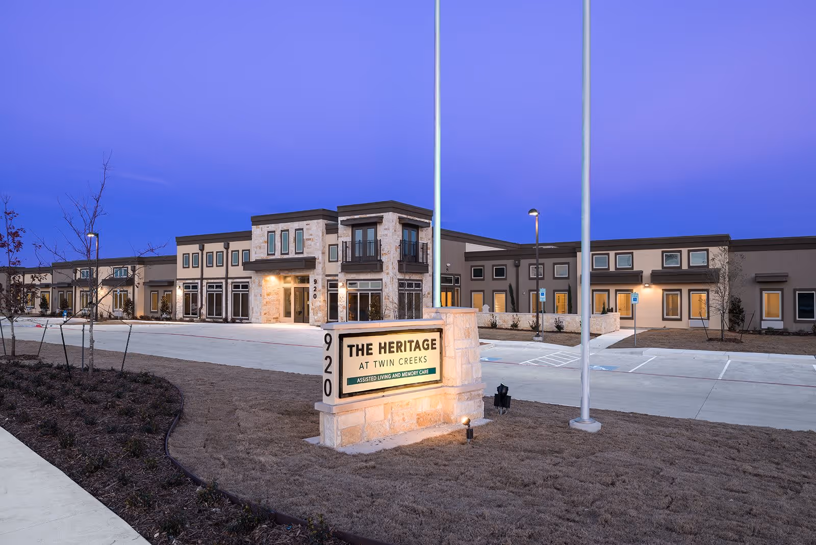Exterior view of The Heritage at Twin Creeks senior living facility at dusk, showing a modern building with multiple windows and a stone entrance. A lit sign in front displays the facility name and indicates it offers assisted living and memory care. The surrounding area includes a paved driveway, parking spaces, and landscaped grounds with young trees and shrubs.