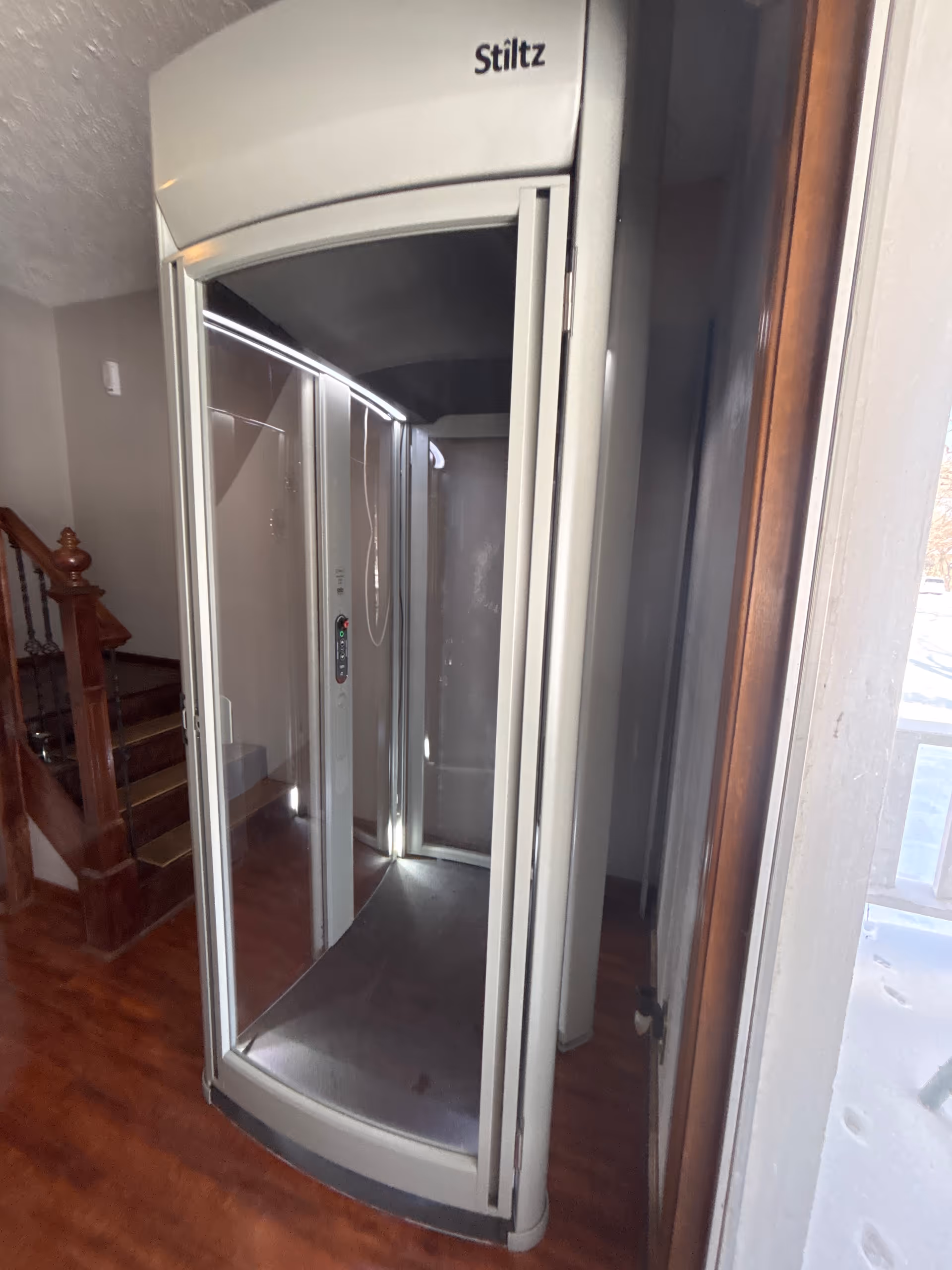 Curved glass elevator in a home interior beside a staircase