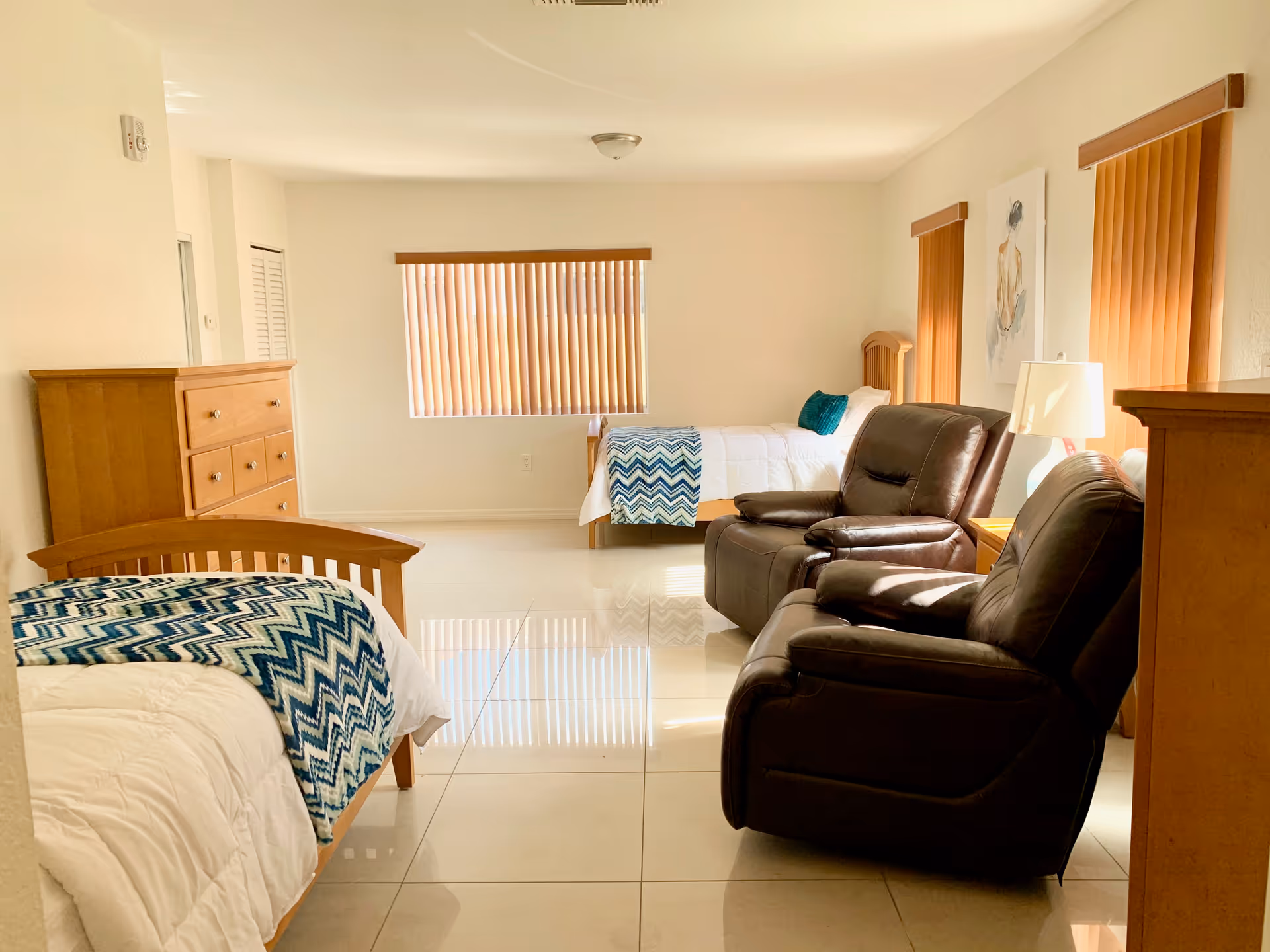 Bright senior living suite with two beds, recliners, dressers, and a window with blinds.