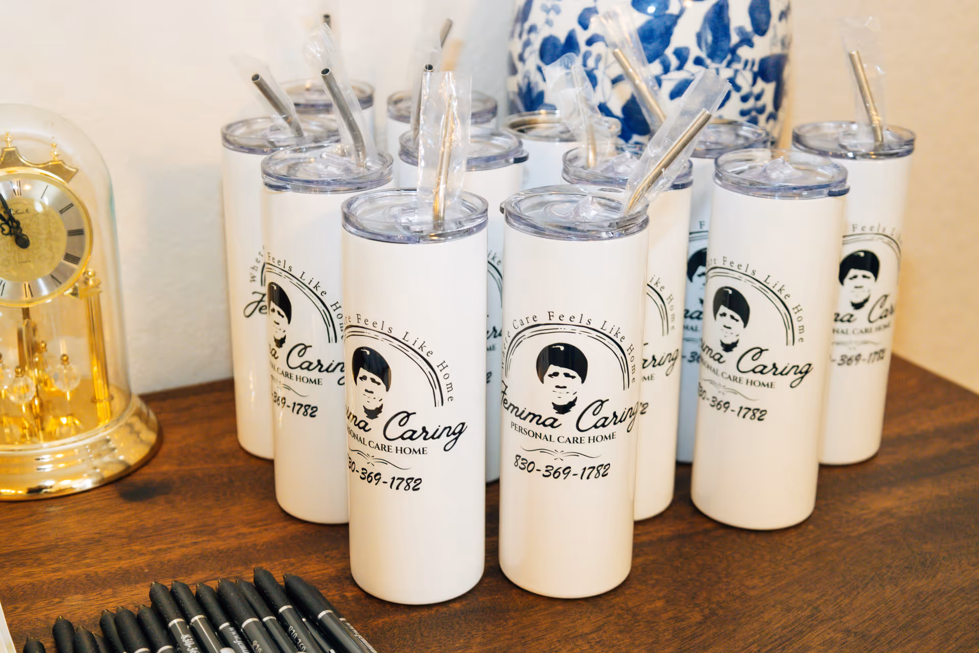 Group of white branded tumblers with straws on a wooden table beside a gold clock.