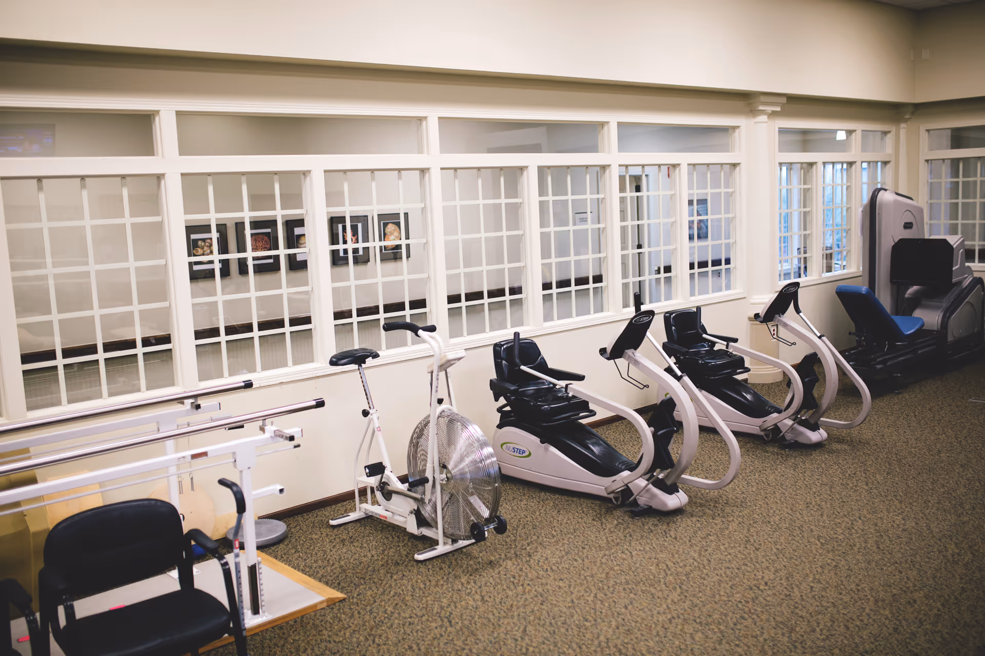 Indoor exercise room with various fitness equipment including stationary bikes, recumbent exercise machines, and parallel bars. The room has beige walls, carpeted floor, and large windows with white grid frames looking into another room with framed pictures on the wall.
