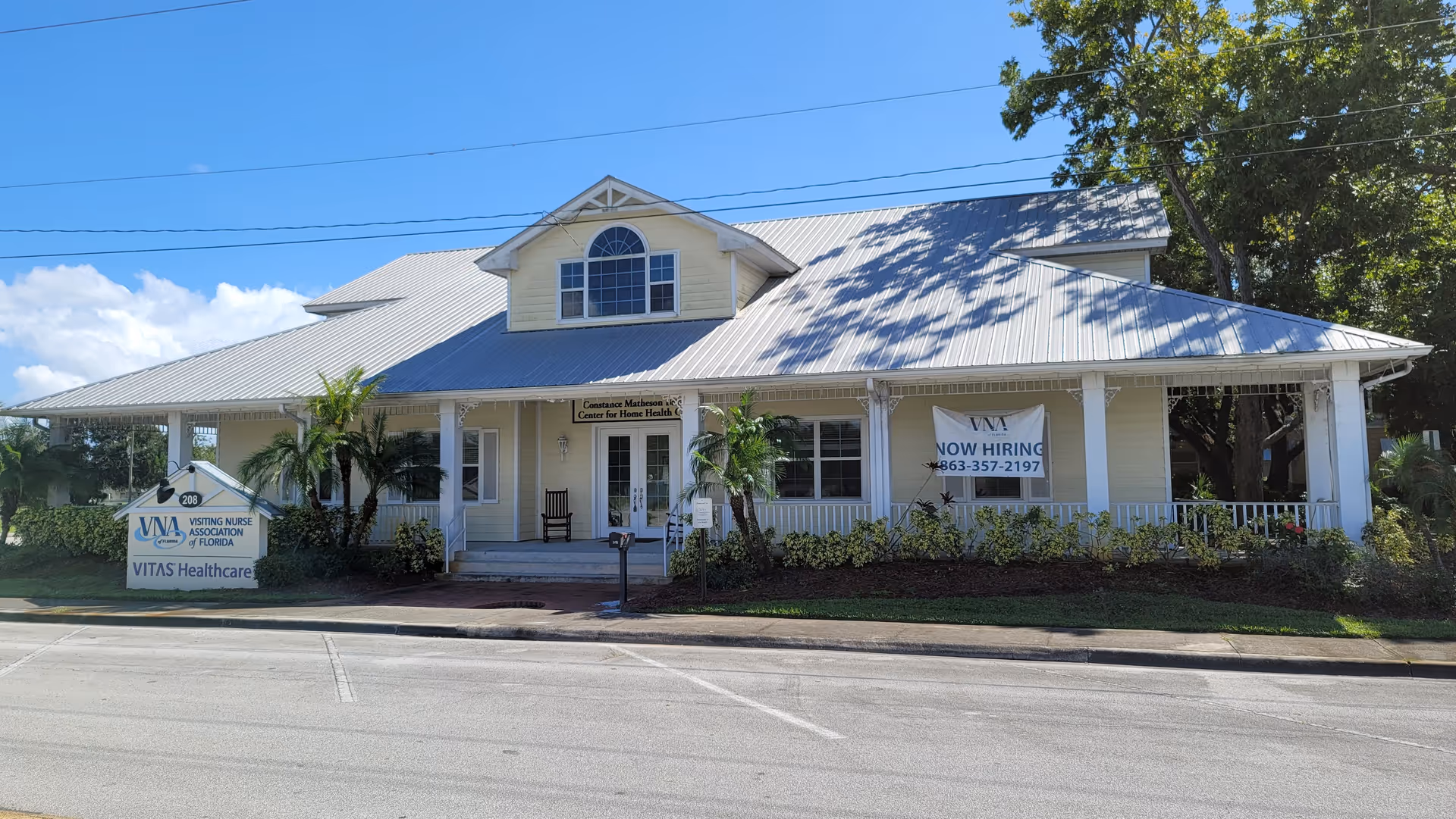 A light-colored, single-story building with a wraparound porch and banners along a street.