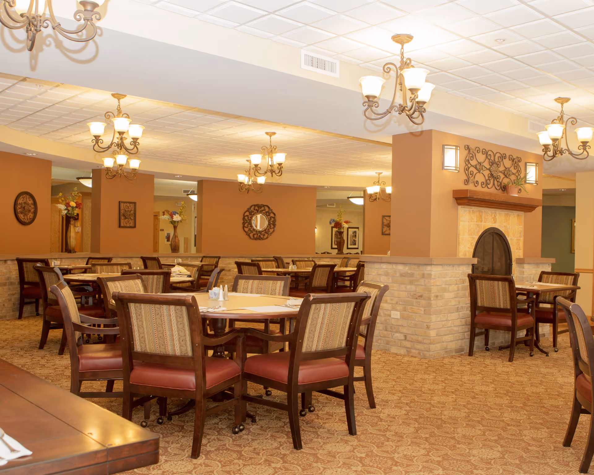 A warm and inviting dining room with multiple round tables and cushioned chairs arranged neatly. The room features beige walls, decorative wall art, a fireplace with a stone surround, and several ornate chandeliers hanging from the ceiling providing soft lighting. The carpet has a patterned design, and there are floral arrangements placed around the room.