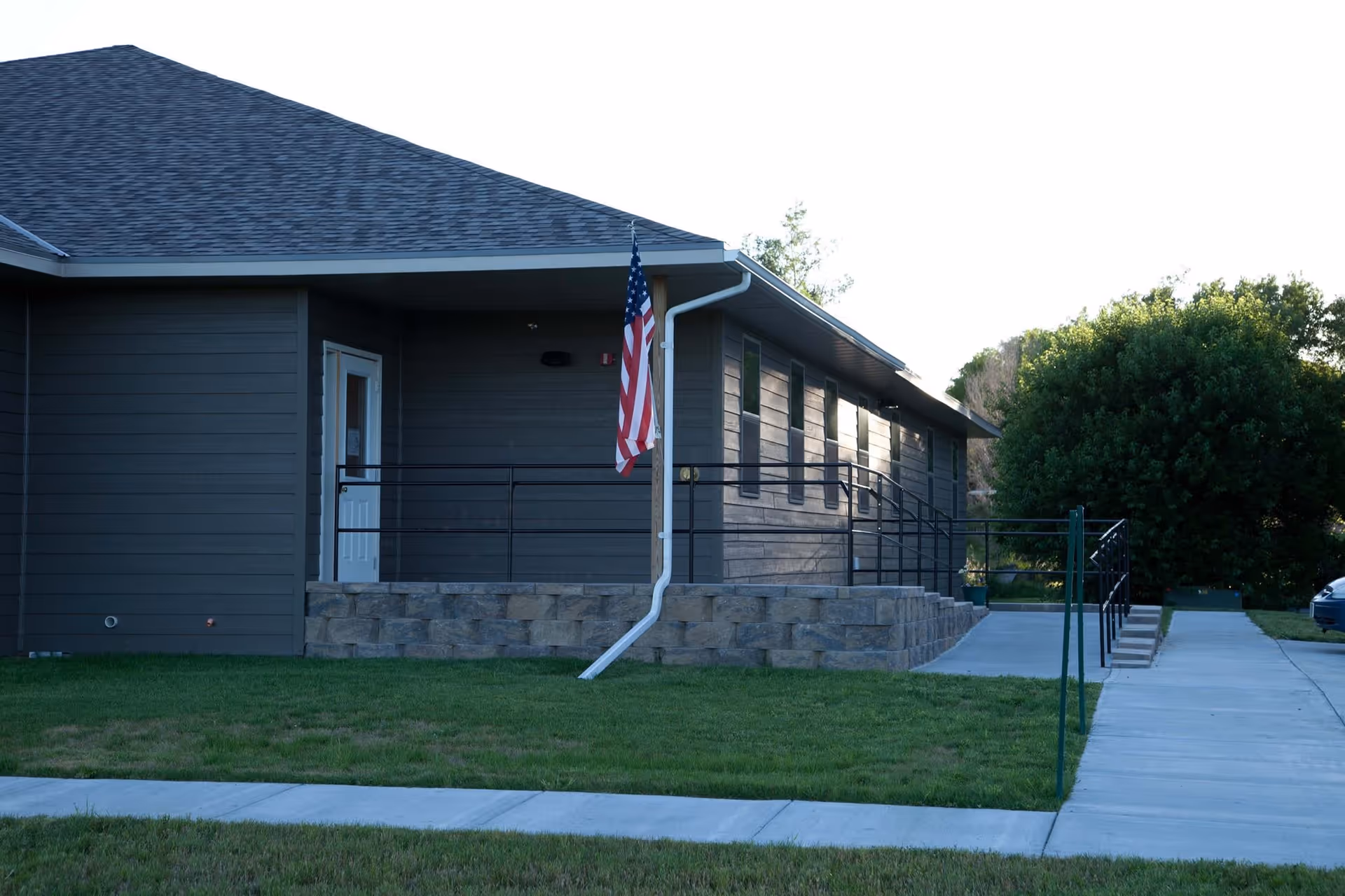 Exterior view of a single-story building with dark siding and a sloped roof. There is a concrete walkway leading to a ramp with black railings and a small set of stairs. An American flag is mounted on a pole near the entrance. Green grass and trees surround the building.
