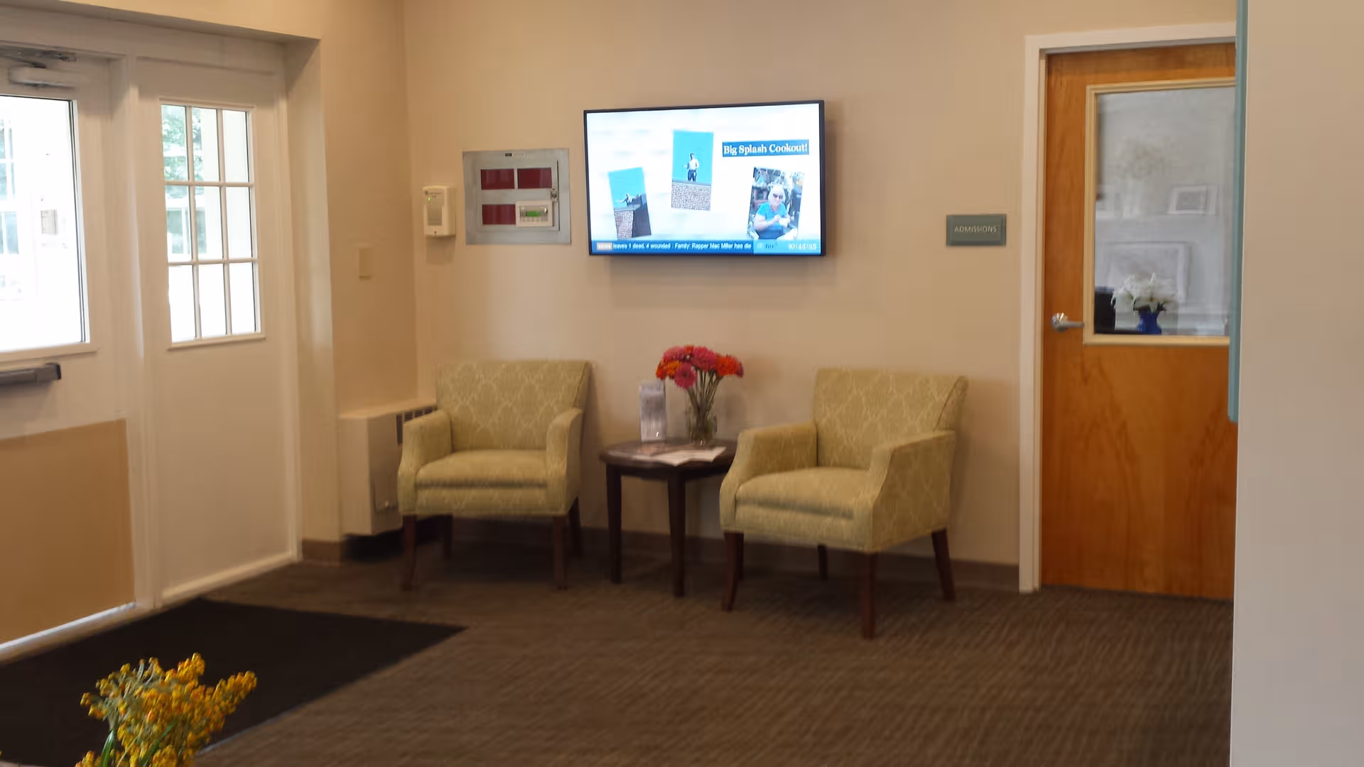 Two beige chairs flank a small table with flowers in a waiting lounge, TV on wall.