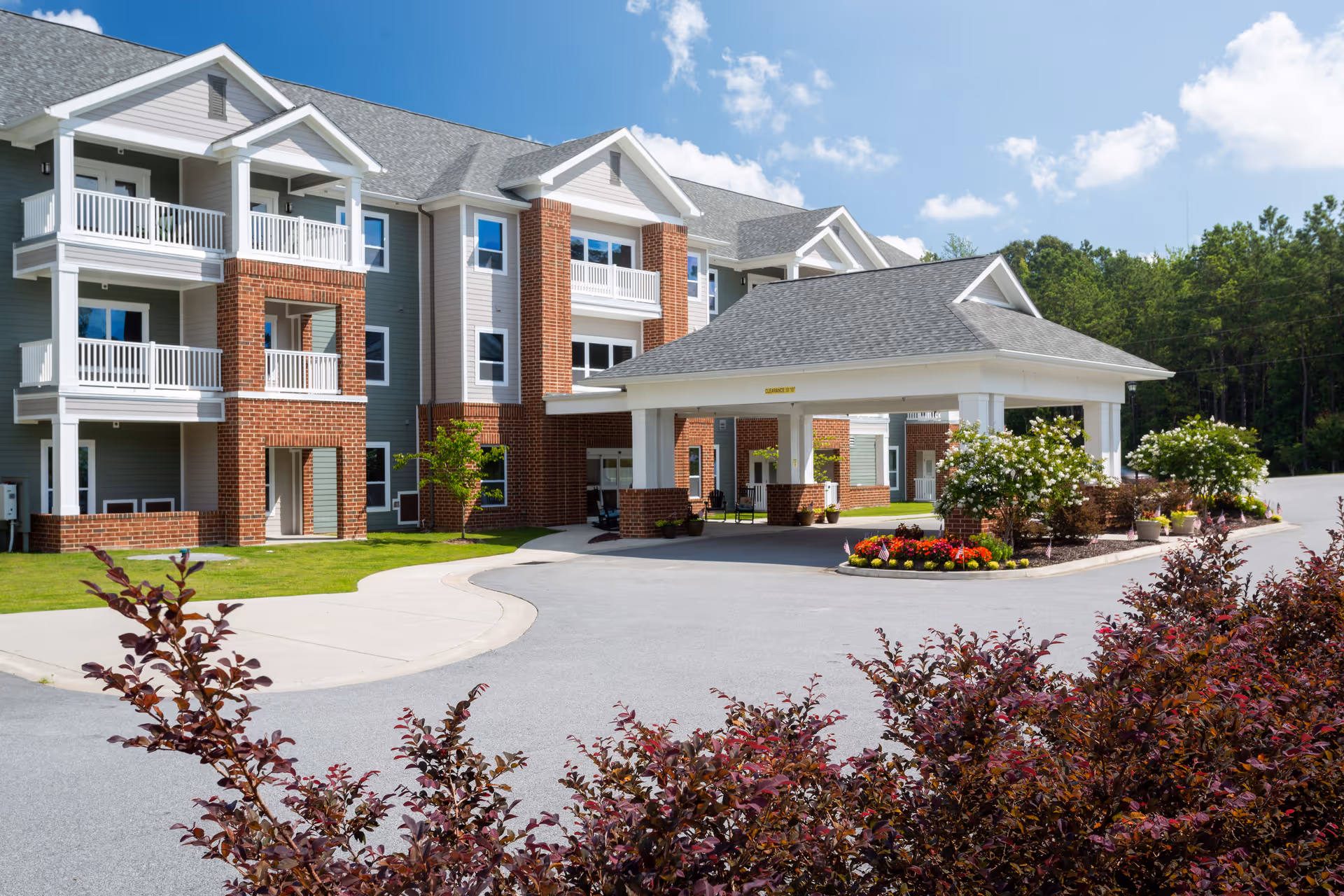 Front exterior of a three-story residential building with white balconies, a covered porte-cochère entrance, and landscaped flower beds.