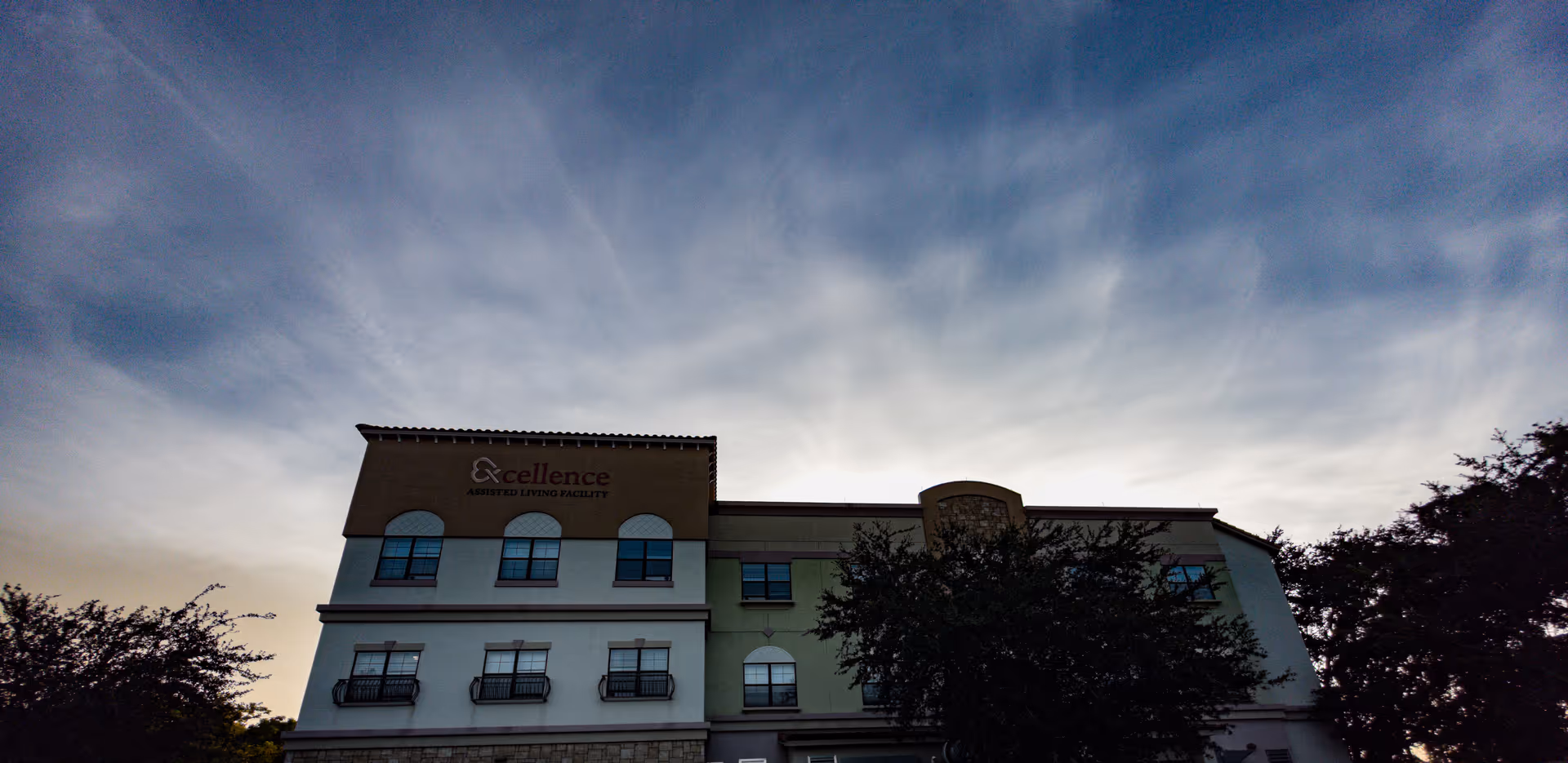 Exterior view of Excellence Senior Living Facility with trees and a bright sky.