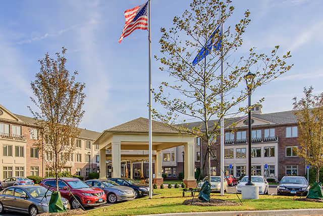 Exterior view of Brookdale Gables Farmington senior living facility showing a parking lot with several cars, a covered entrance with columns, two flagpoles with the American flag and another flag, and trees with green leaves under a partly cloudy sky.