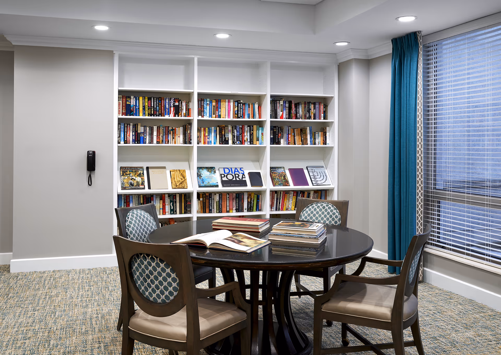 Round table with open books, four cushioned chairs, and tall bookshelves in a library room.