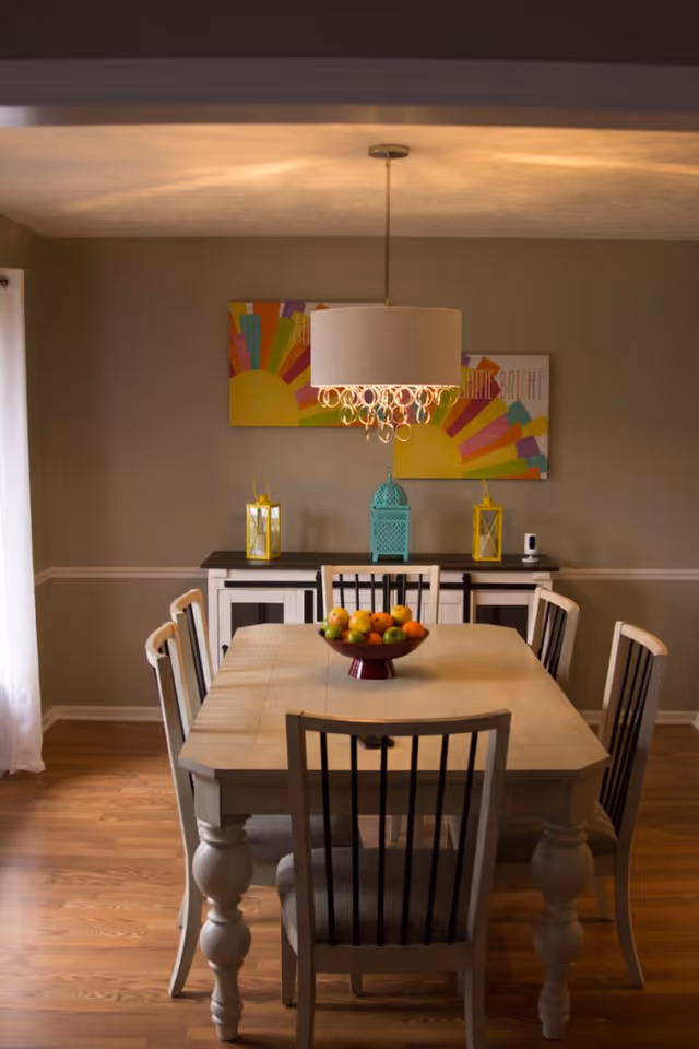 A dining room with a wooden table and six chairs. On the table is a bowl filled with various fruits. Above the table hangs a modern chandelier with a white shade and decorative hanging crystals. Against the back wall is a sideboard with two yellow lanterns and a turquoise decorative lantern. Two colorful paintings featuring a sunburst design are mounted on the wall above the sideboard. The room has wooden flooring and neutral-colored walls with white trim.