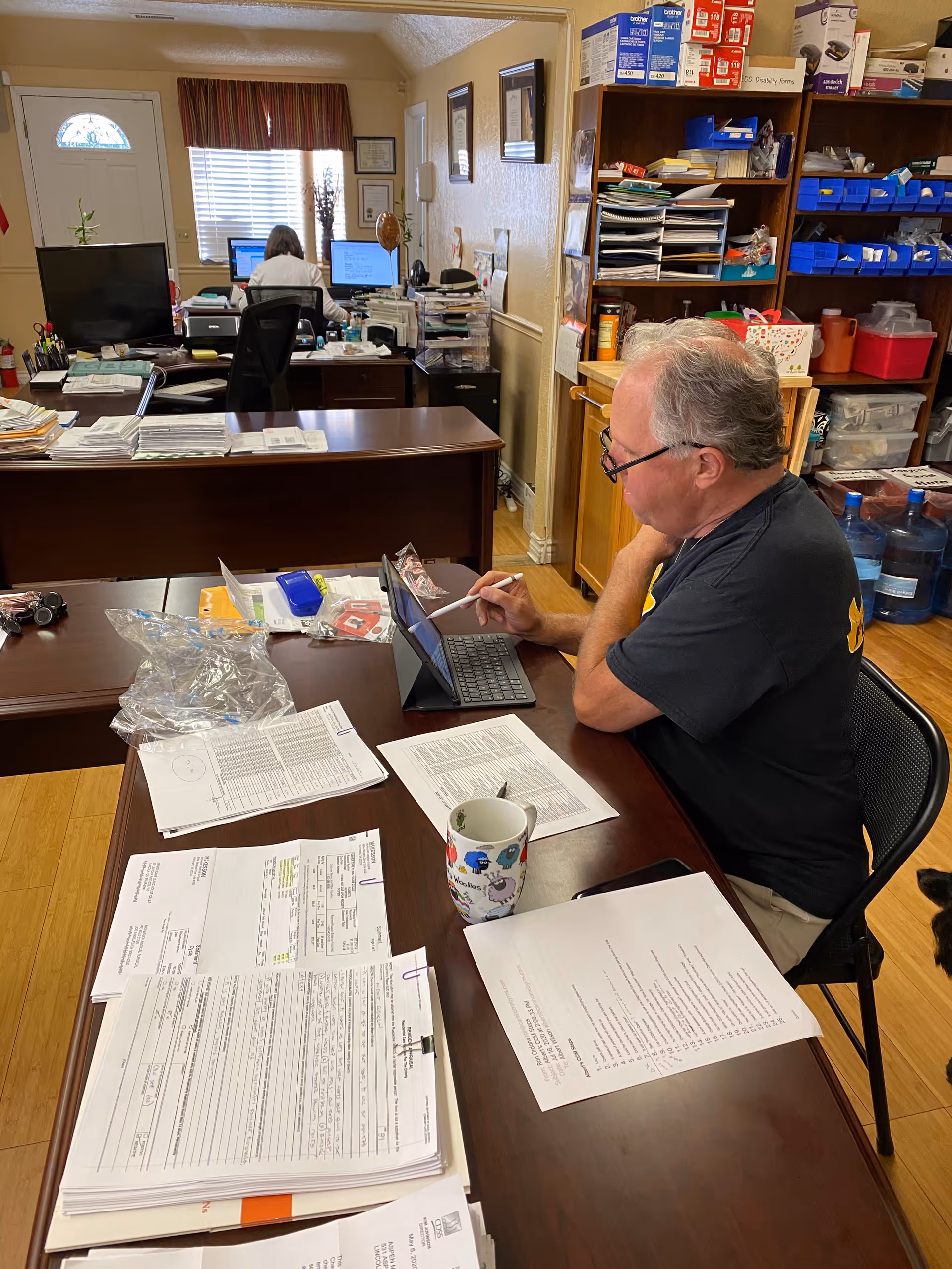 An office setting with two people working at desks. A man is seated at a table in the foreground, using a tablet with a stylus, surrounded by papers and a colorful mug. In the background, a woman is working at a computer desk near a window with blinds and a curtain. Shelves with office supplies and water bottles are visible on the right side.