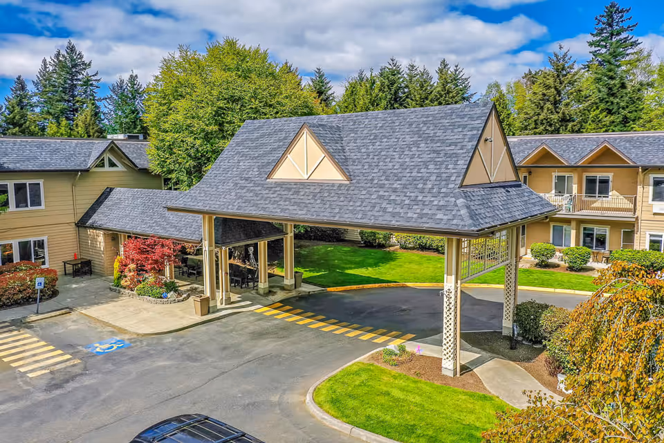 Exterior view of Solstice Senior Living At Normandy Park showing a covered entrance with a peaked roof, surrounded by a driveway, green lawns, trees, and a two-story building with beige siding and multiple windows.