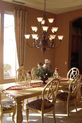 A dining room with a wooden table and six chairs featuring patterned cushions. The table is decorated with a red and gold runner, a floral centerpiece, and two candle holders with red candles. A large chandelier with multiple lights hangs above the table. There is a window with beige curtains allowing natural light into the room.