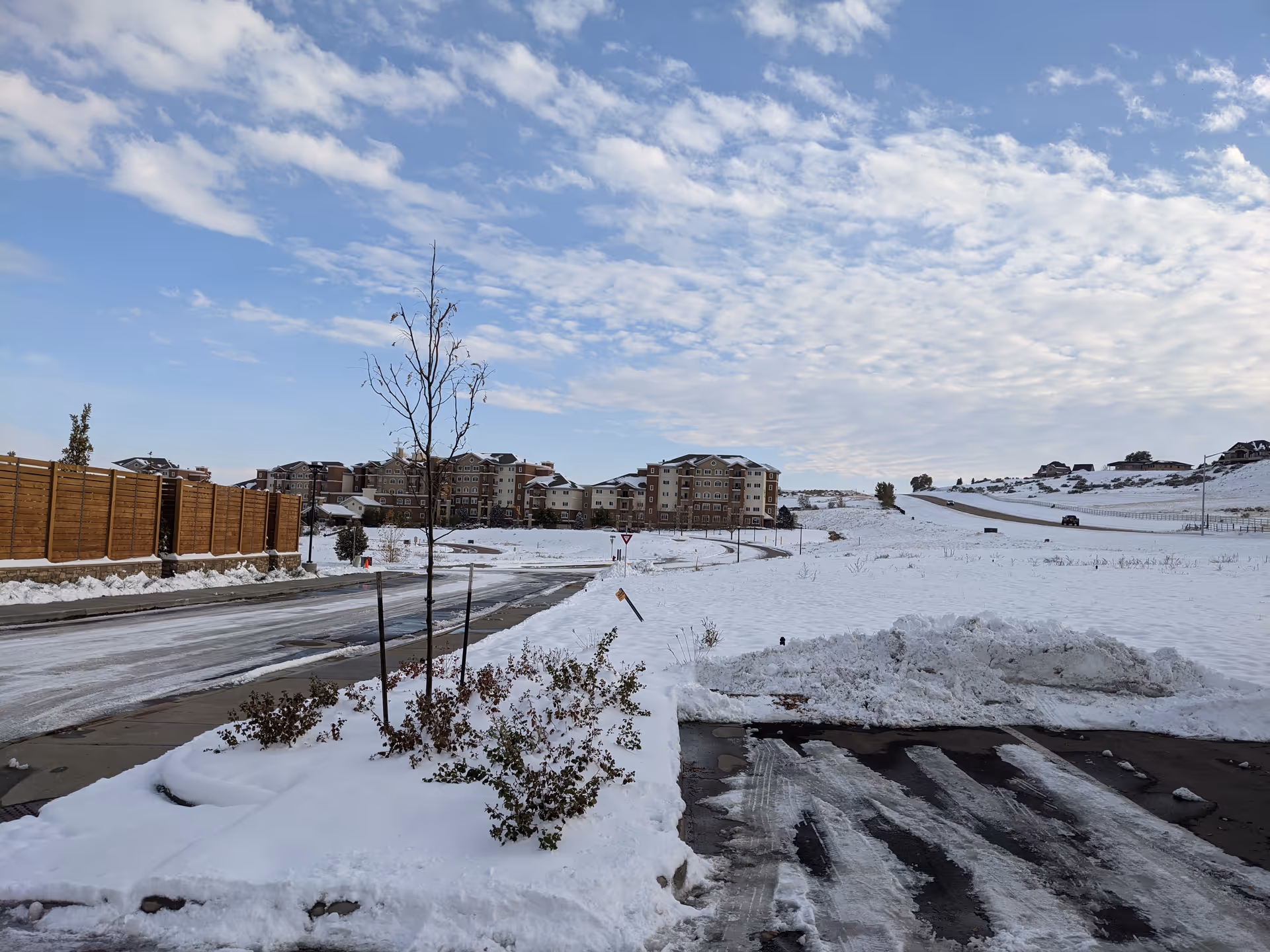 Snow-covered landscape with a road, wooden fence, and distant residential buildings.