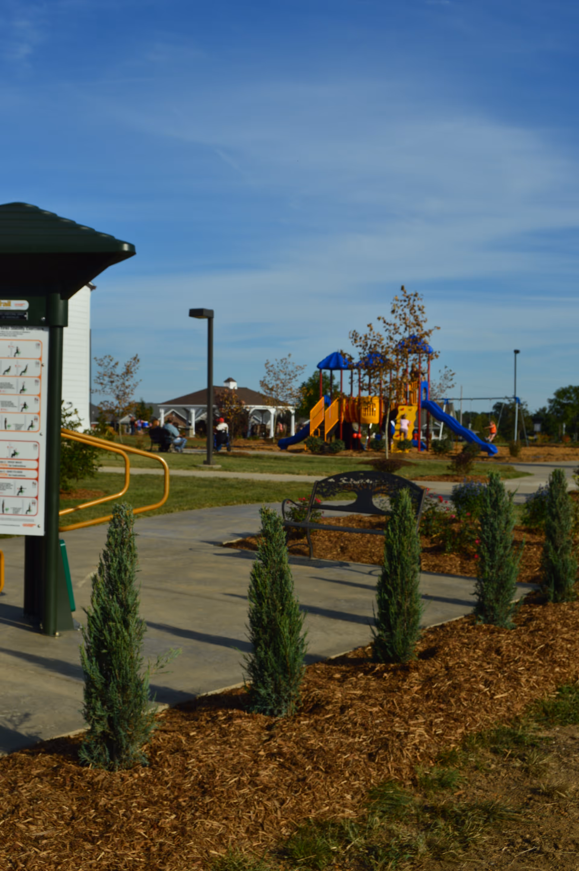 Outdoor area of Magnolia Terrace Senior Living Apartments featuring a paved walkway lined with small evergreen shrubs, a bench, a playground with slides and climbing structures, and a gazebo in the background under a blue sky.