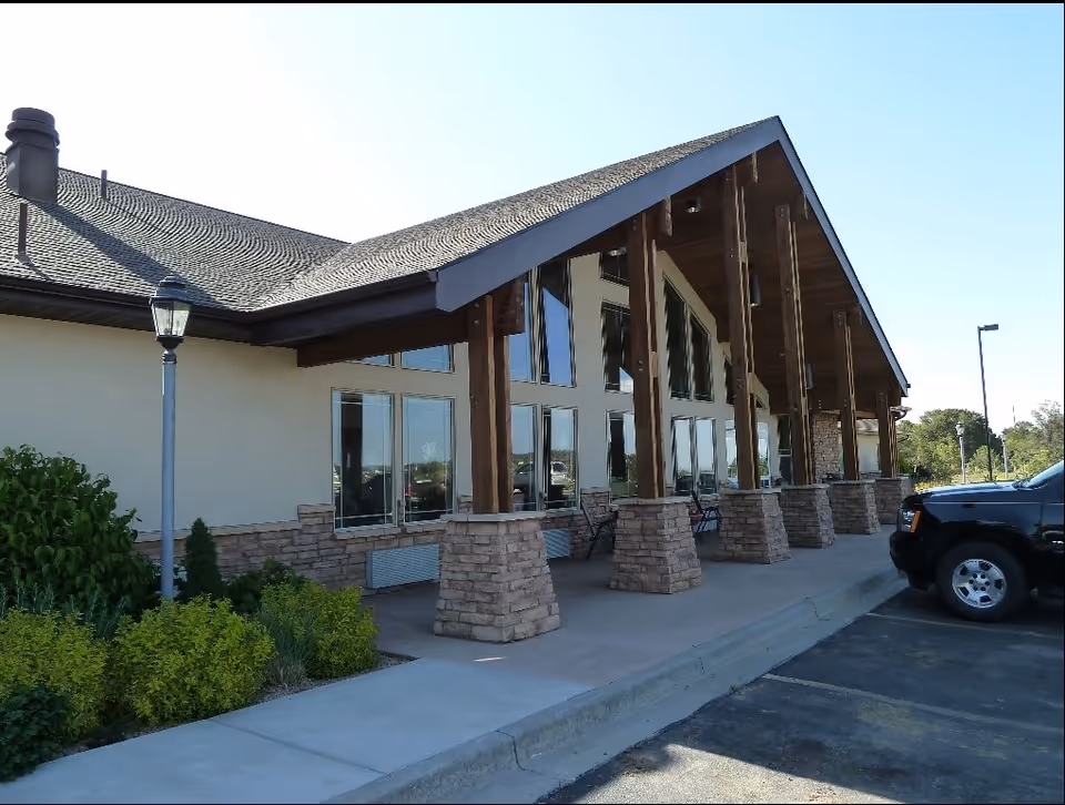 Exterior view of a building with a sloped roof supported by wooden beams on stone bases. There are large windows along the wall and a sidewalk with some greenery and a lamp post. A black vehicle is parked on the right side.