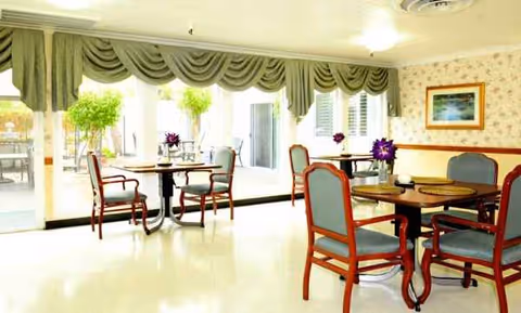 Bright dining room with round tables, cushioned chairs, and glass doors in a senior facility.