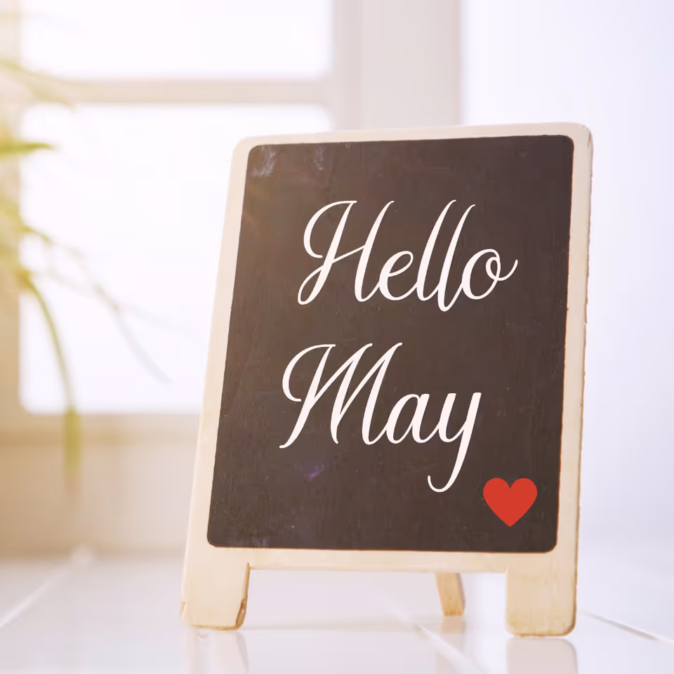 Chalkboard sign reading Hello May with a small red heart on a bright indoor surface.