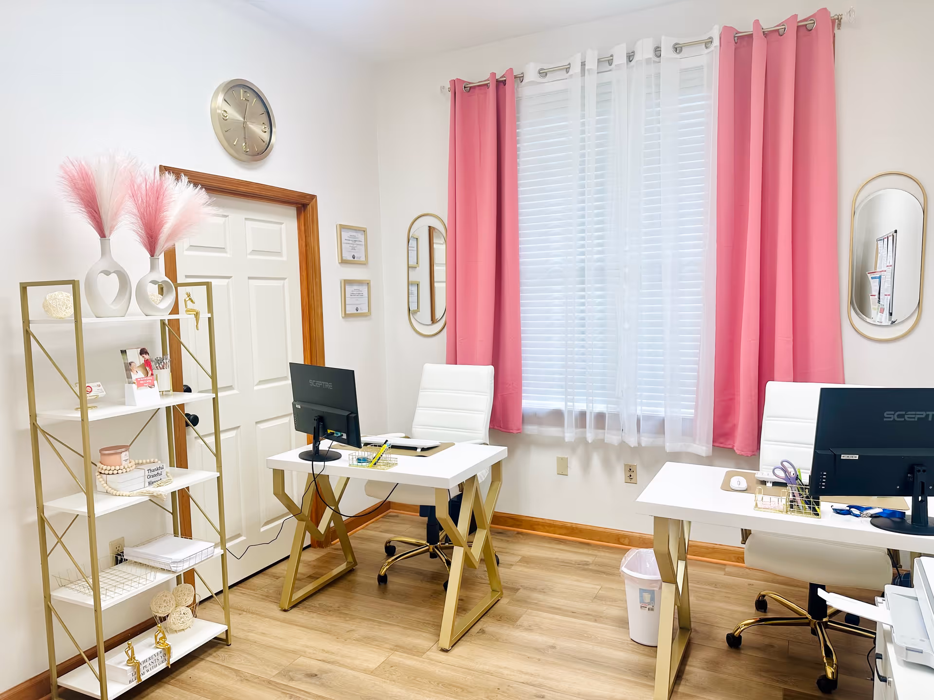 A bright office area with two white desks, computers, pink curtains, and decorative shelves.