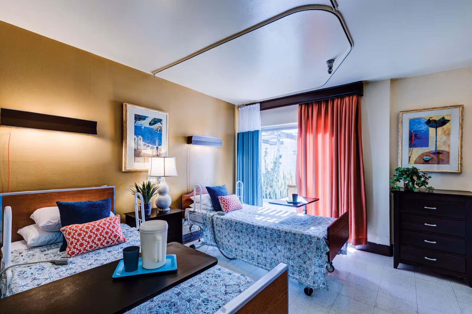Two hospital-style beds in a bright patient room with curtains, furniture, and artwork.